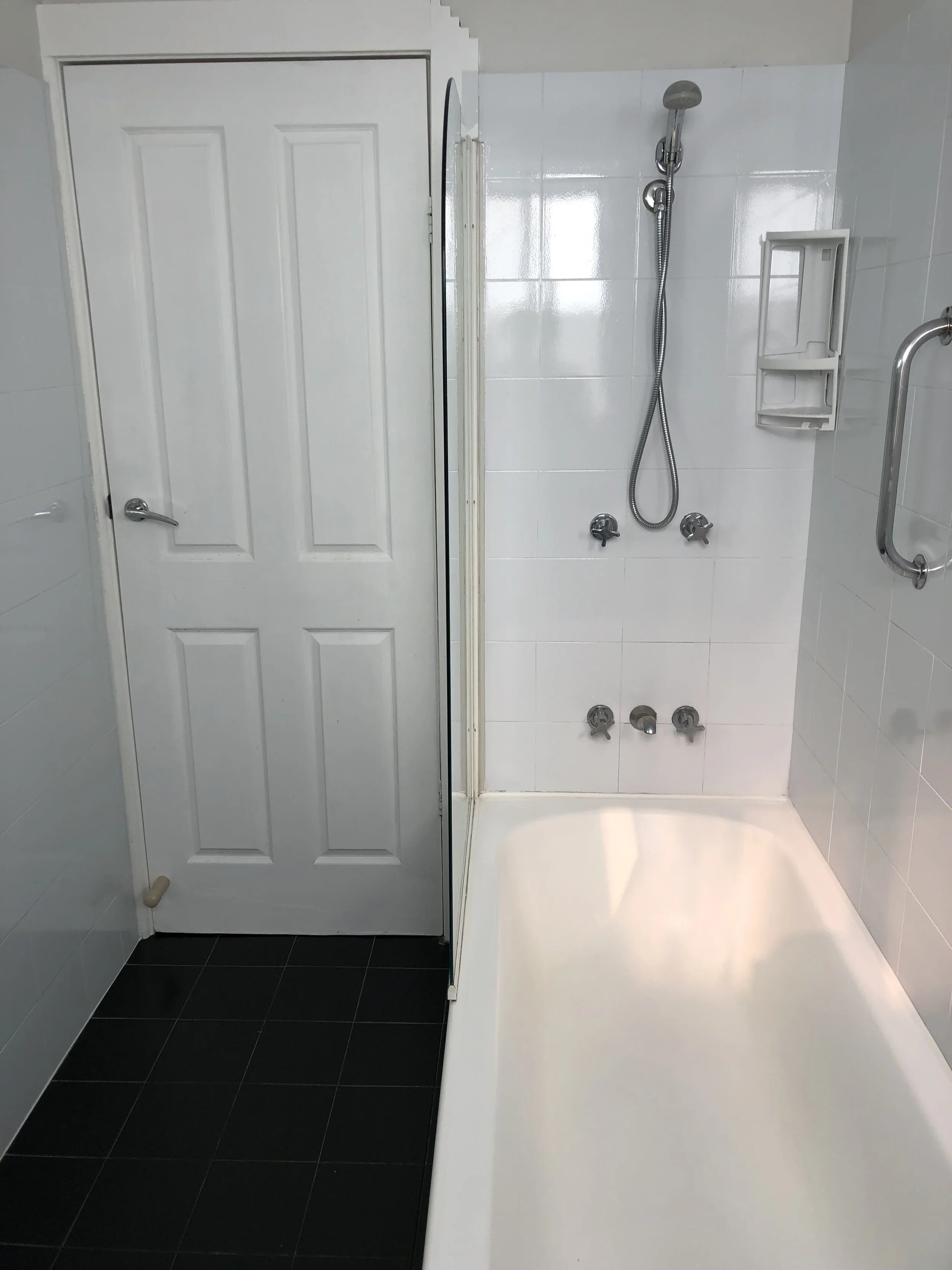 A small shower area with a white tiled wall, a handheld showerhead, and four control knobs, next to a closed white door and a black tiled floor.