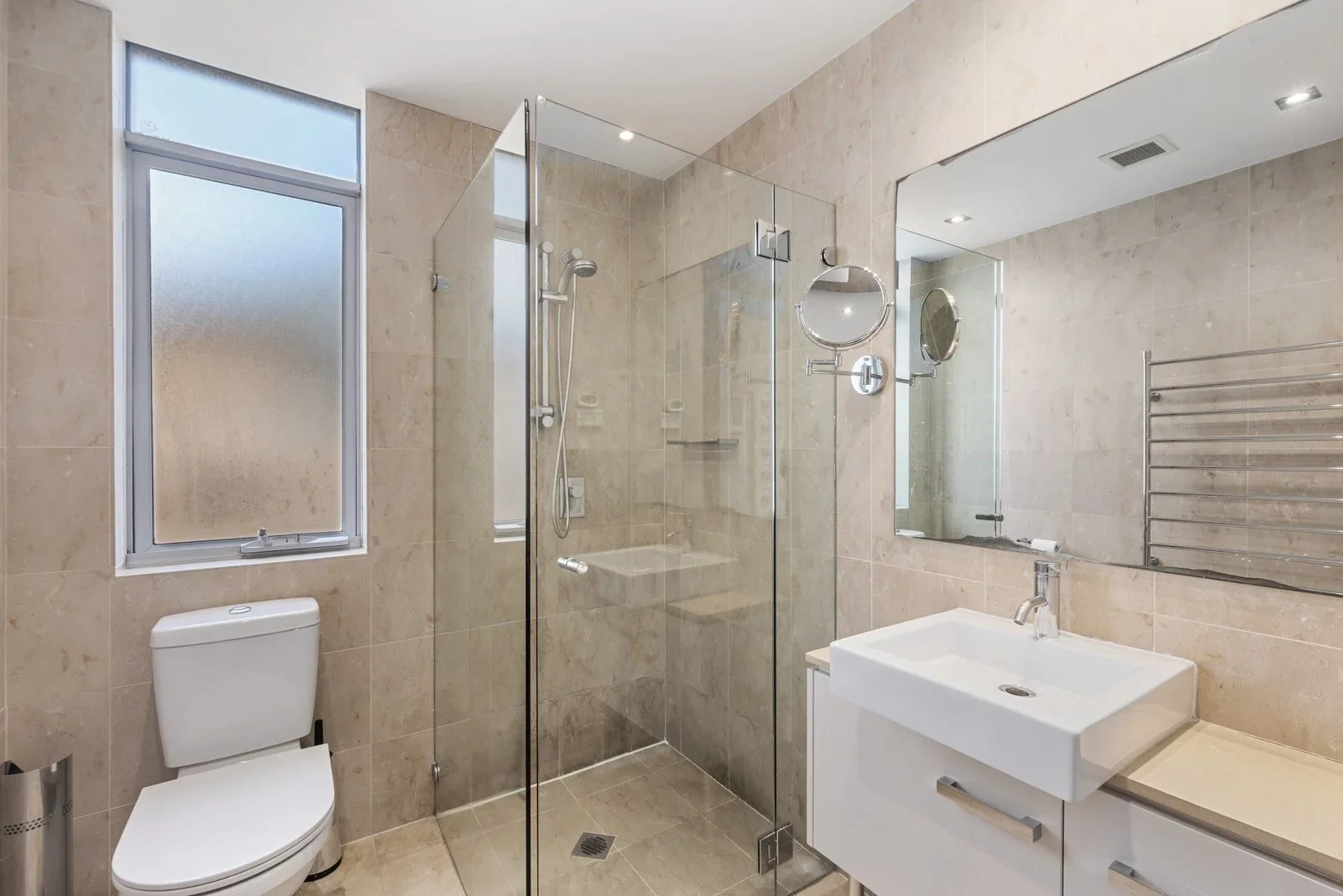 Modern bathroom with beige tiled walls and floor, glass shower enclosure with chrome fixtures, white toilet, and white vanity with a rectangular sink. A large mirror and a heated towel rail are also visible.