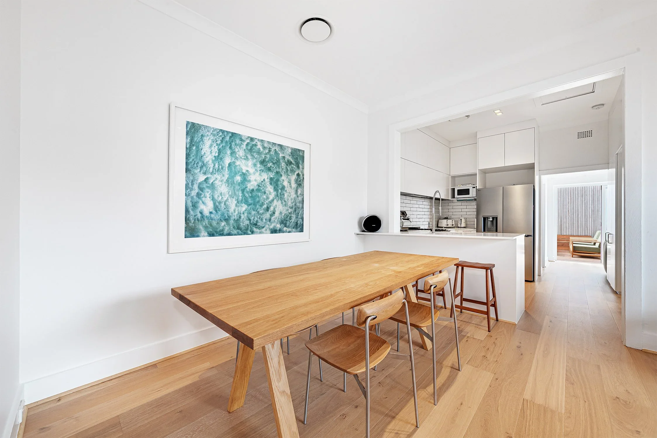 Kitchen with white cabinets, appliances, and a wooden dining table with four chairs, next to a white wall with a large abstract artwork.