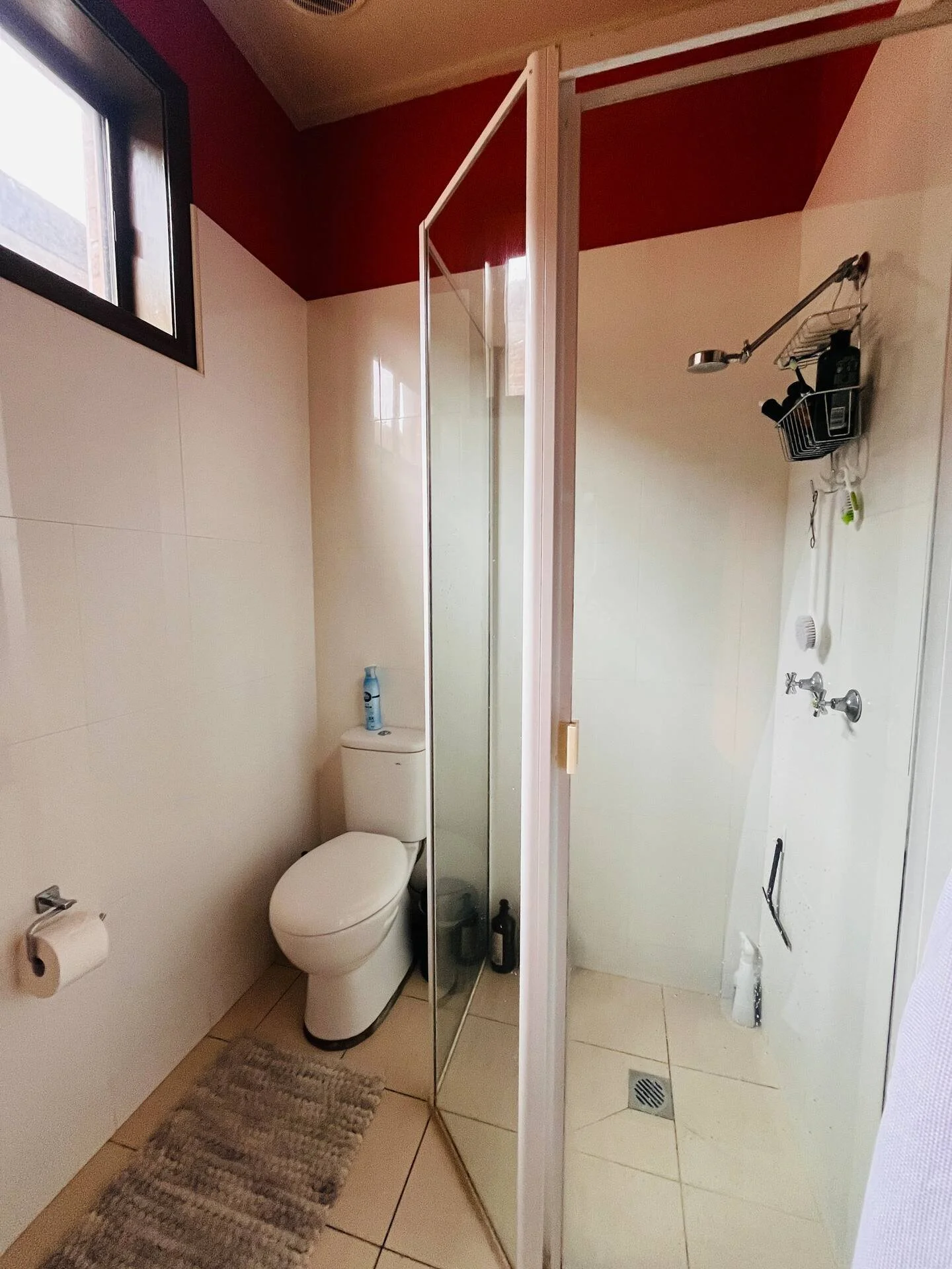 Small bathroom with a toilet, a shower with a glass door, beige tiled floor, beige and red walls, a window, and a small shelf in the shower area.