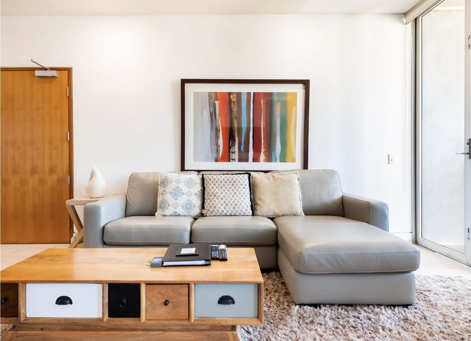 Living room with a beige sectional sofa, four decorative throw pillows, a wooden coffee table with drawers and books, a white rug, abstract art on the wall, a door to the left, a large window to the right.