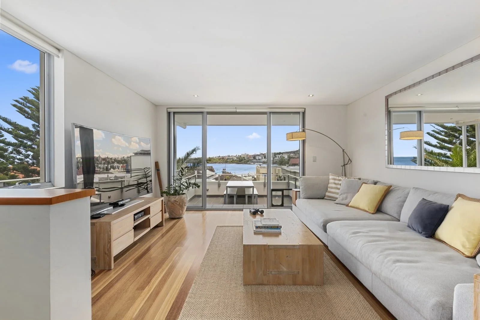 Living room with large sliding glass doors leading to balcony, view of water and cityscape, light-colored sofa with pillows, wooden coffee table, wall mirror, television, potted plant, floor lamp, and windows with ocean view.
