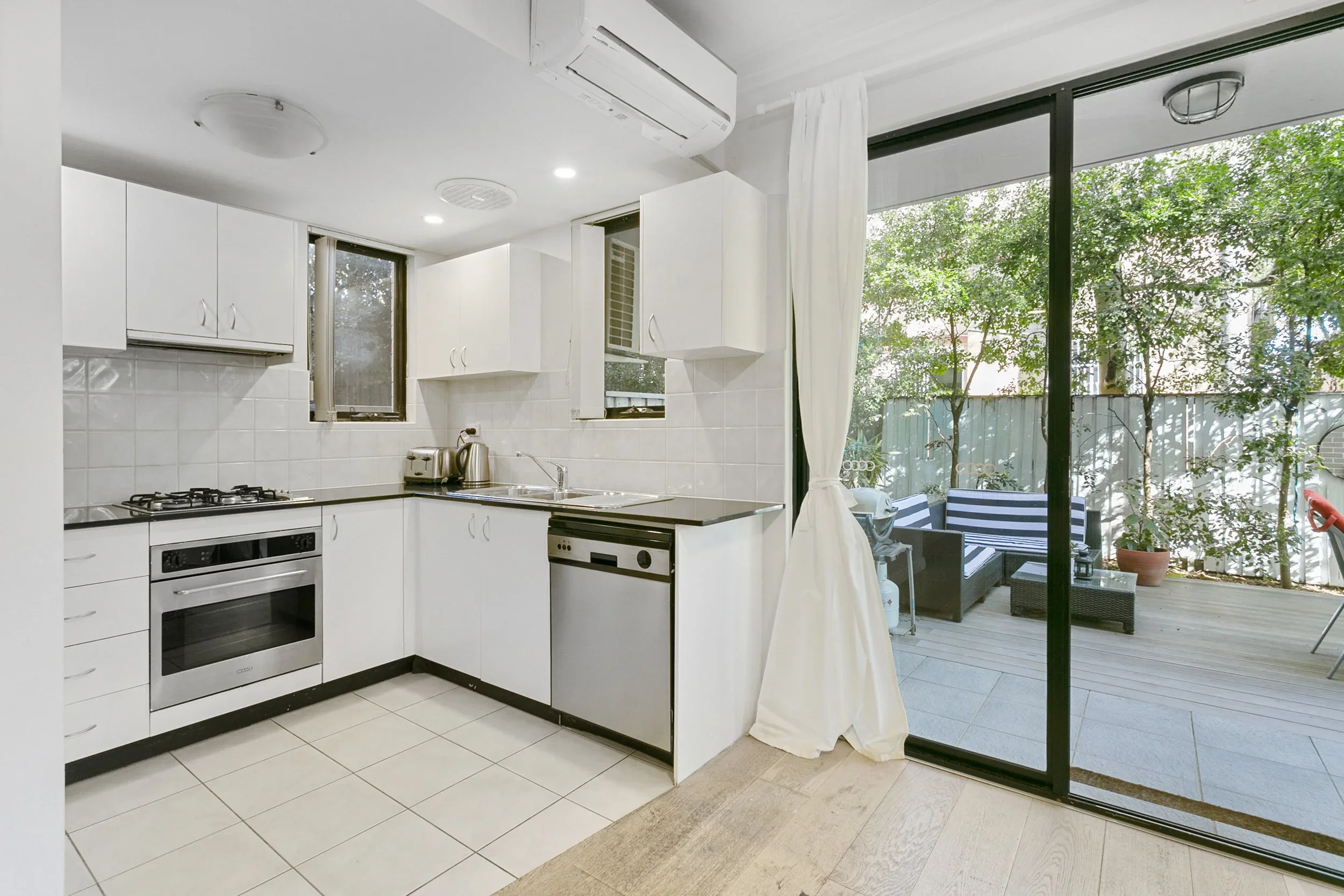 Kitchen with white cabinets and a sliding glass door leading to an outdoor patio with seating and greenery.