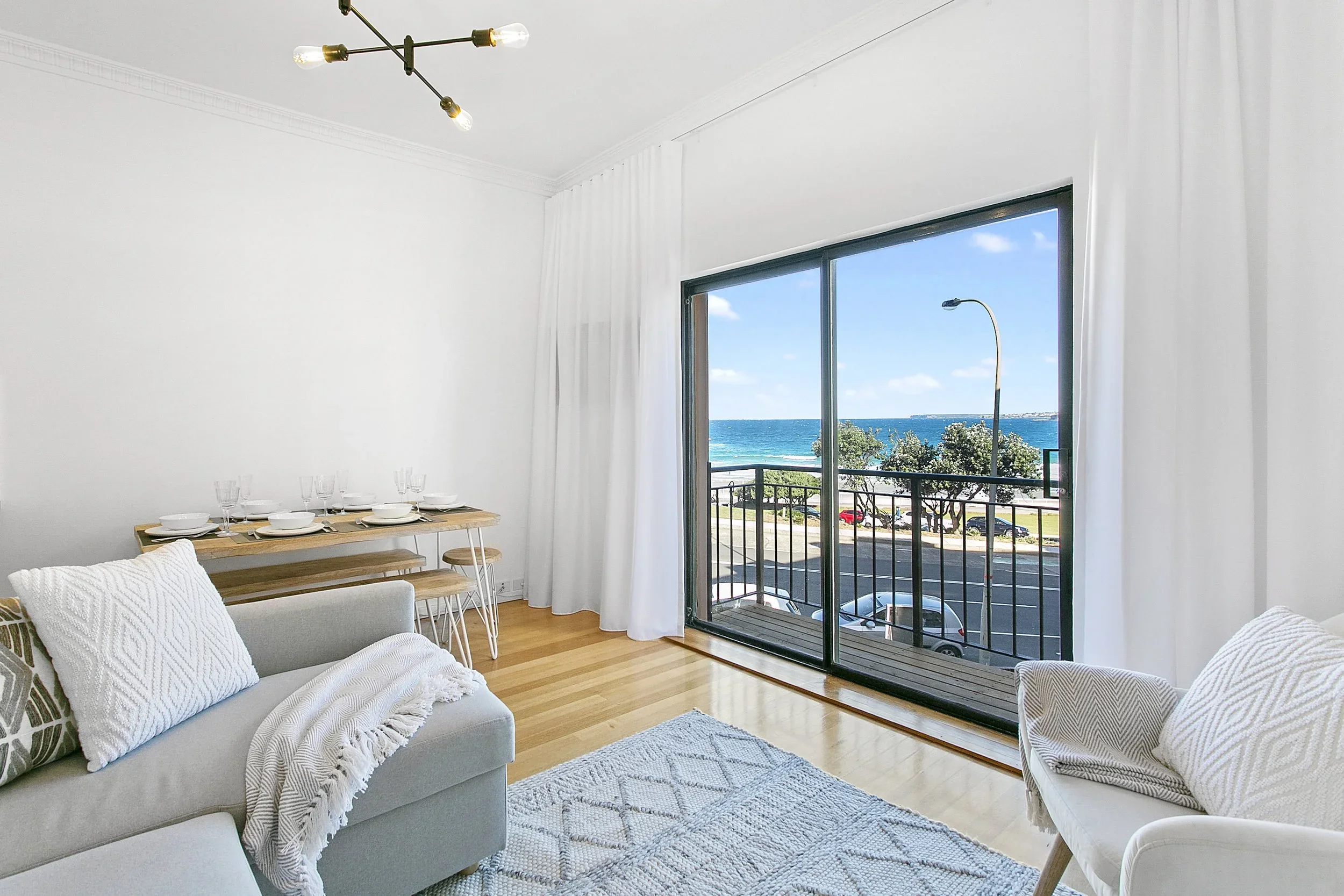 Living room with a large sliding glass door overlooking the beach and ocean, decorated with white curtains, a beige sofa with pillows and throw blanket, a wooden dining table with dinnerware, a rug, and a modern chandelier.