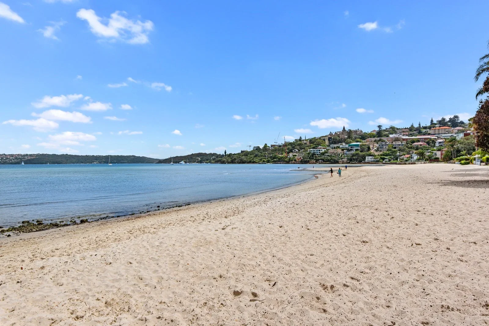 A sandy beach along the water with a few people walking, calm blue water, and houses on a hillside in the background under a partly cloudy sky.
