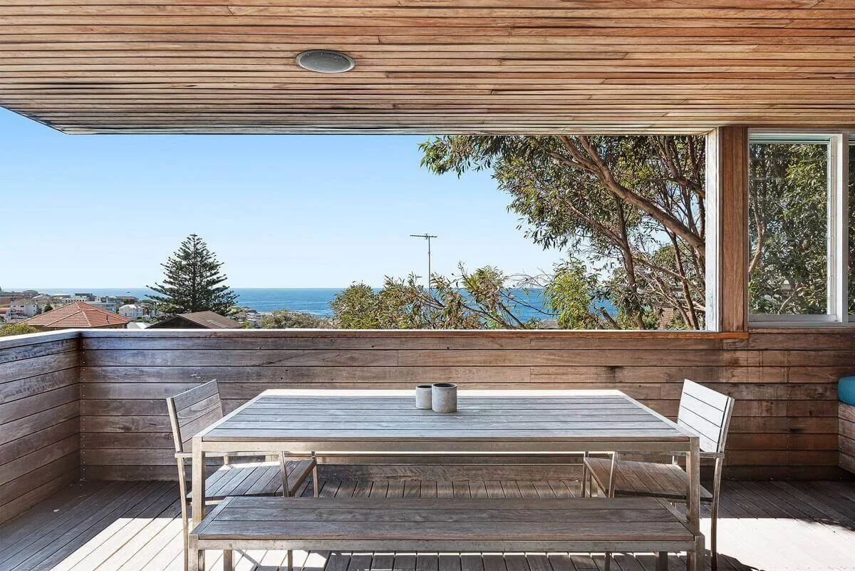 Balcony with wooden furniture and a view of trees, houses, and the ocean in the distance under a clear sky.