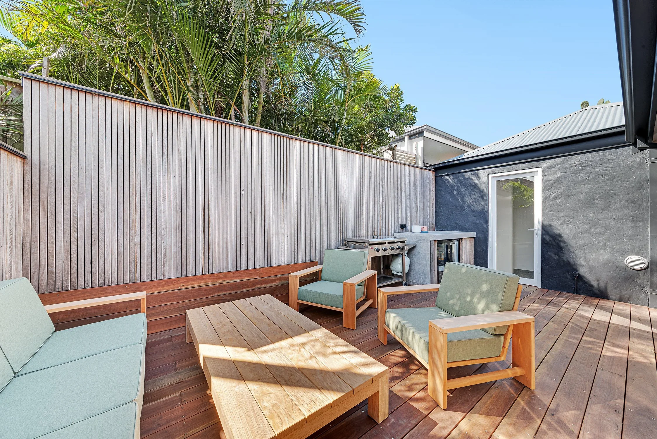 Outdoor patio with wooden deck furniture including a sofa, two armchairs, and a coffee table, with a charcoal wall, door, and a built-in outdoor kitchen area featuring a grill and sink, surrounded by tall palm trees and a clear blue sky.