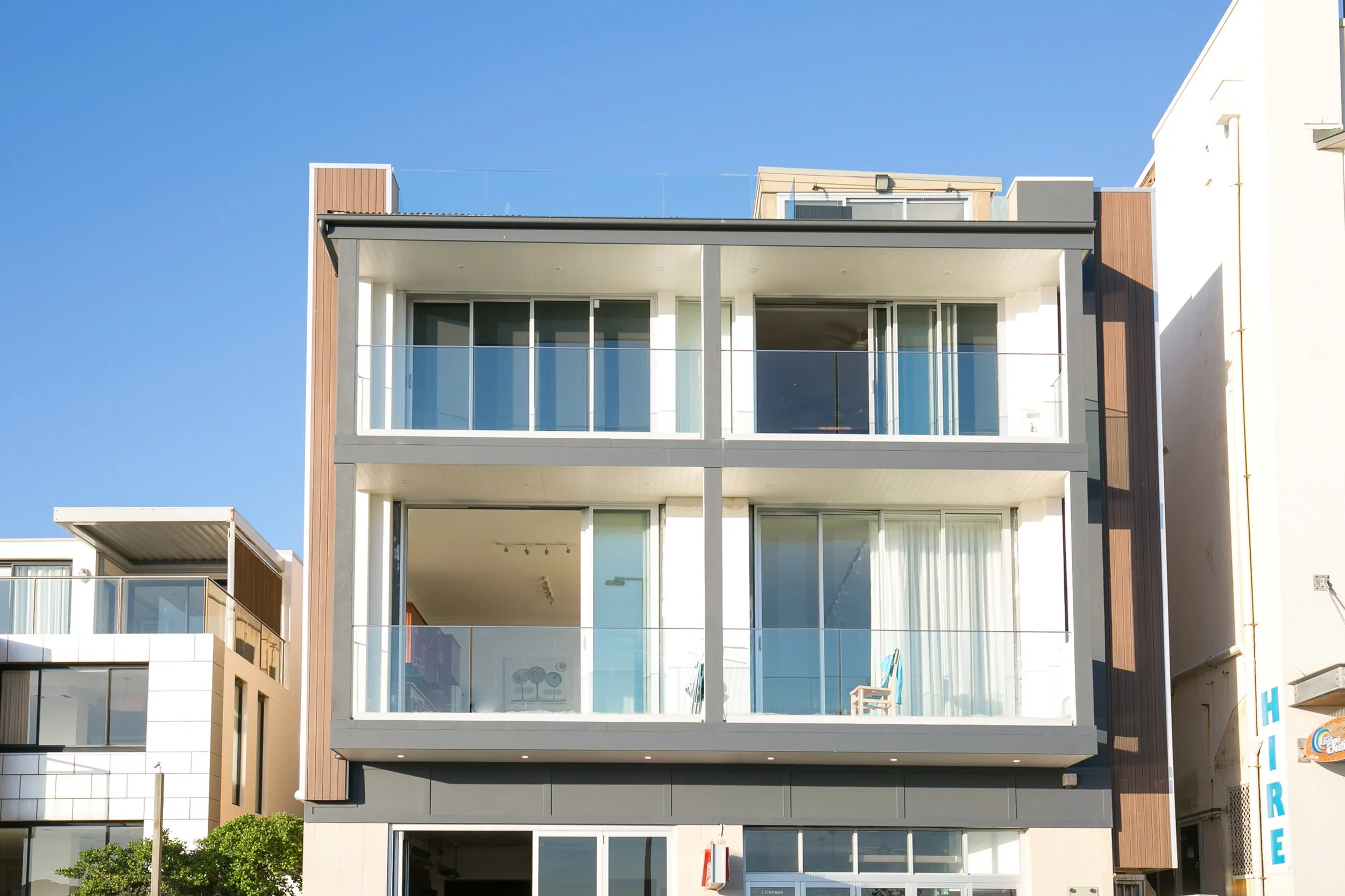 Modern multi-story apartment building with glass balconies and large windows, situated in an urban area under a clear blue sky.