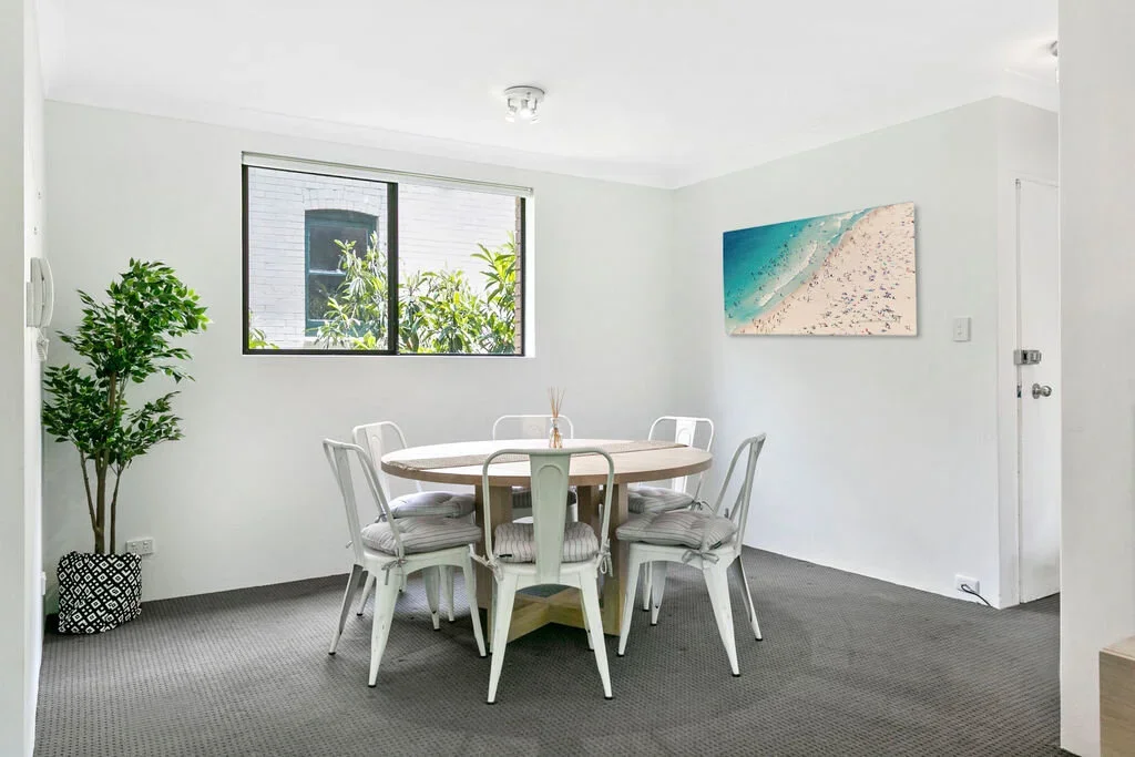 Dining room with a round table, six white chairs with cushions, a potted plant, a window showing greenery outside, and a wall art piece depicting a beach scene.