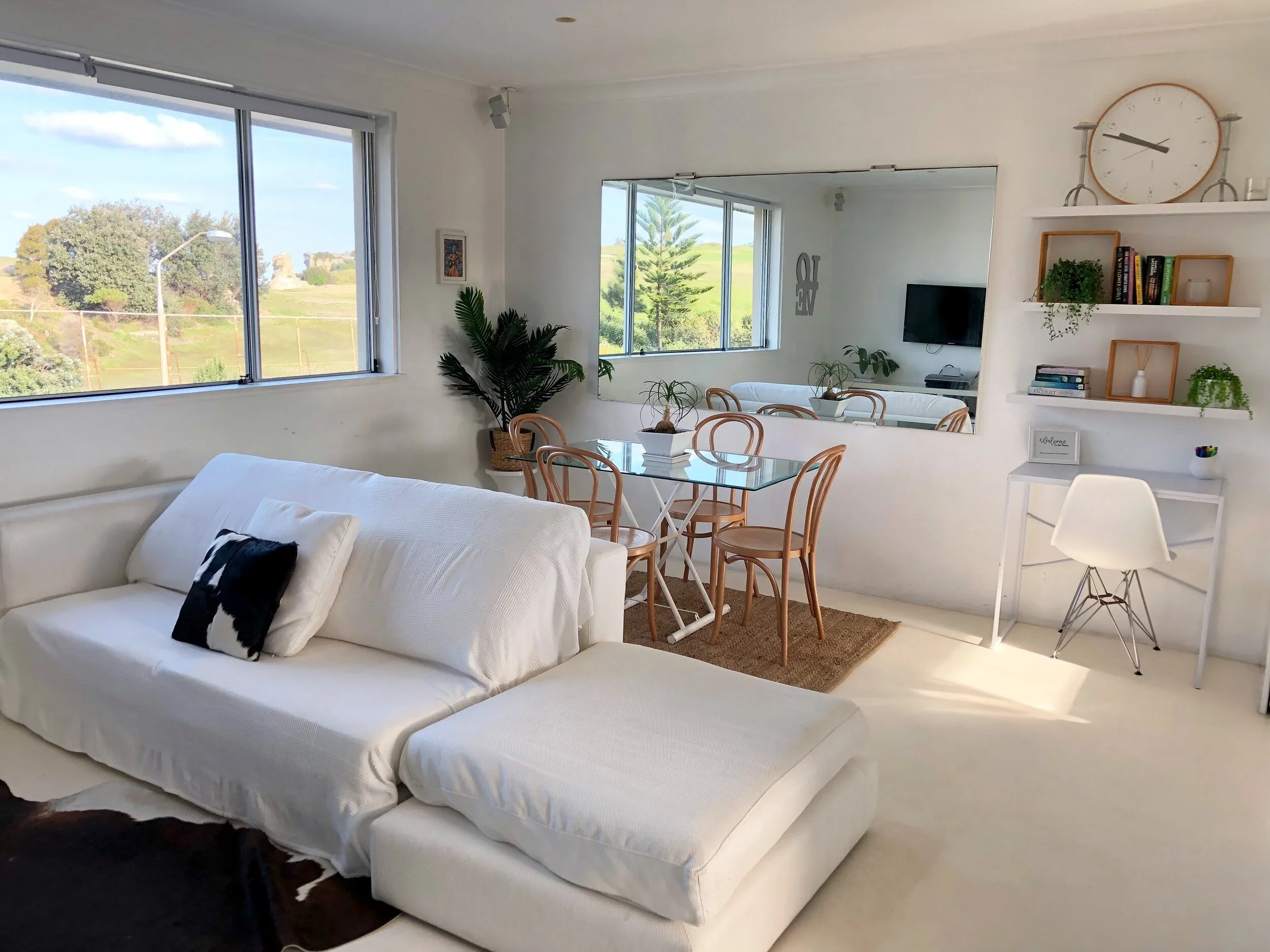 Bright living room with white sofa, glass dining table with wooden chairs, large window showing outdoor scenery, wall mirror, and decorative shelves with books and plants.