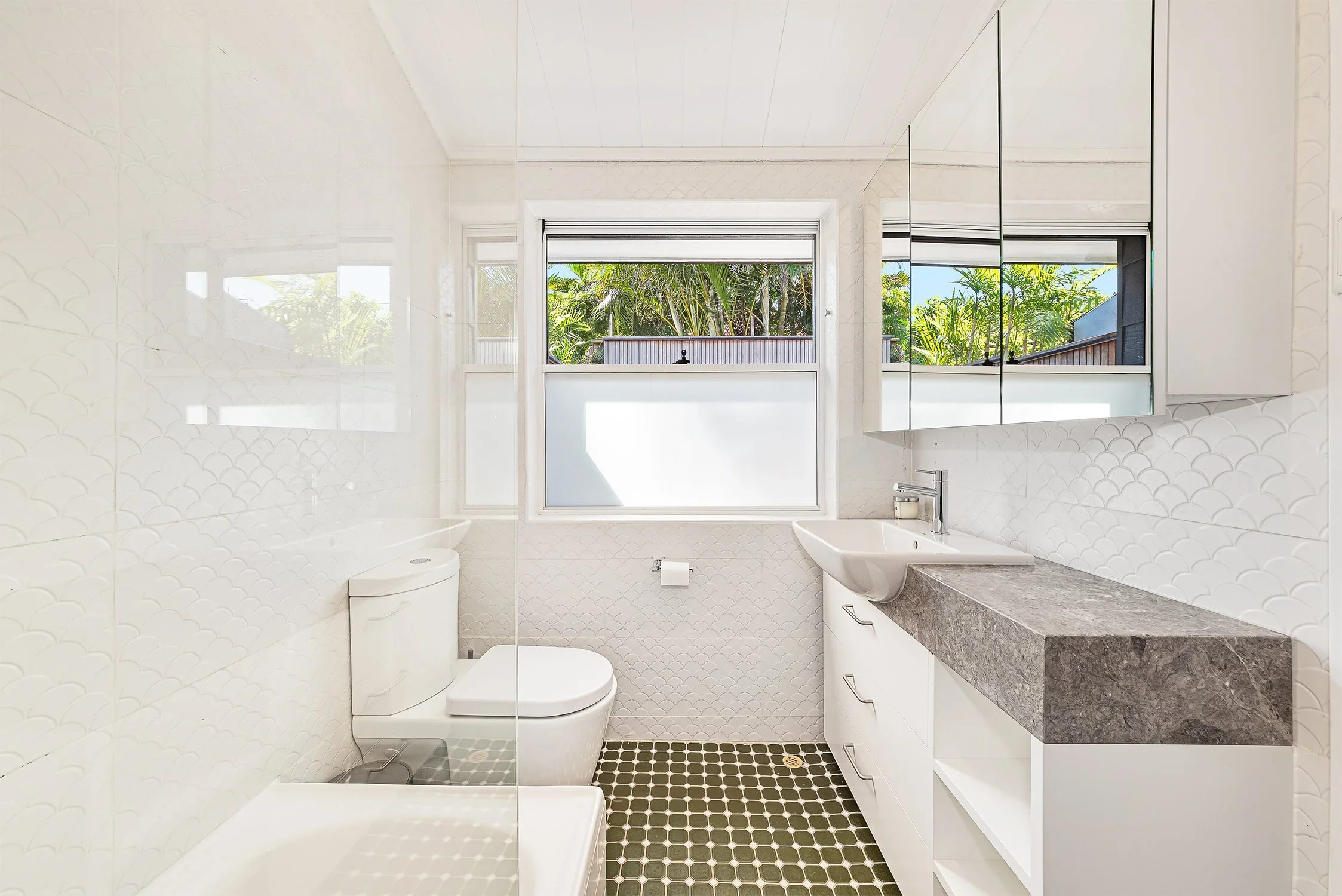 Bright modern bathroom with white walls, a window displaying greenery outside, a white toilet, a white sink with a metallic faucet, and a mirrored cabinet above the sink. The floor has small, dark green hexagon tiles.