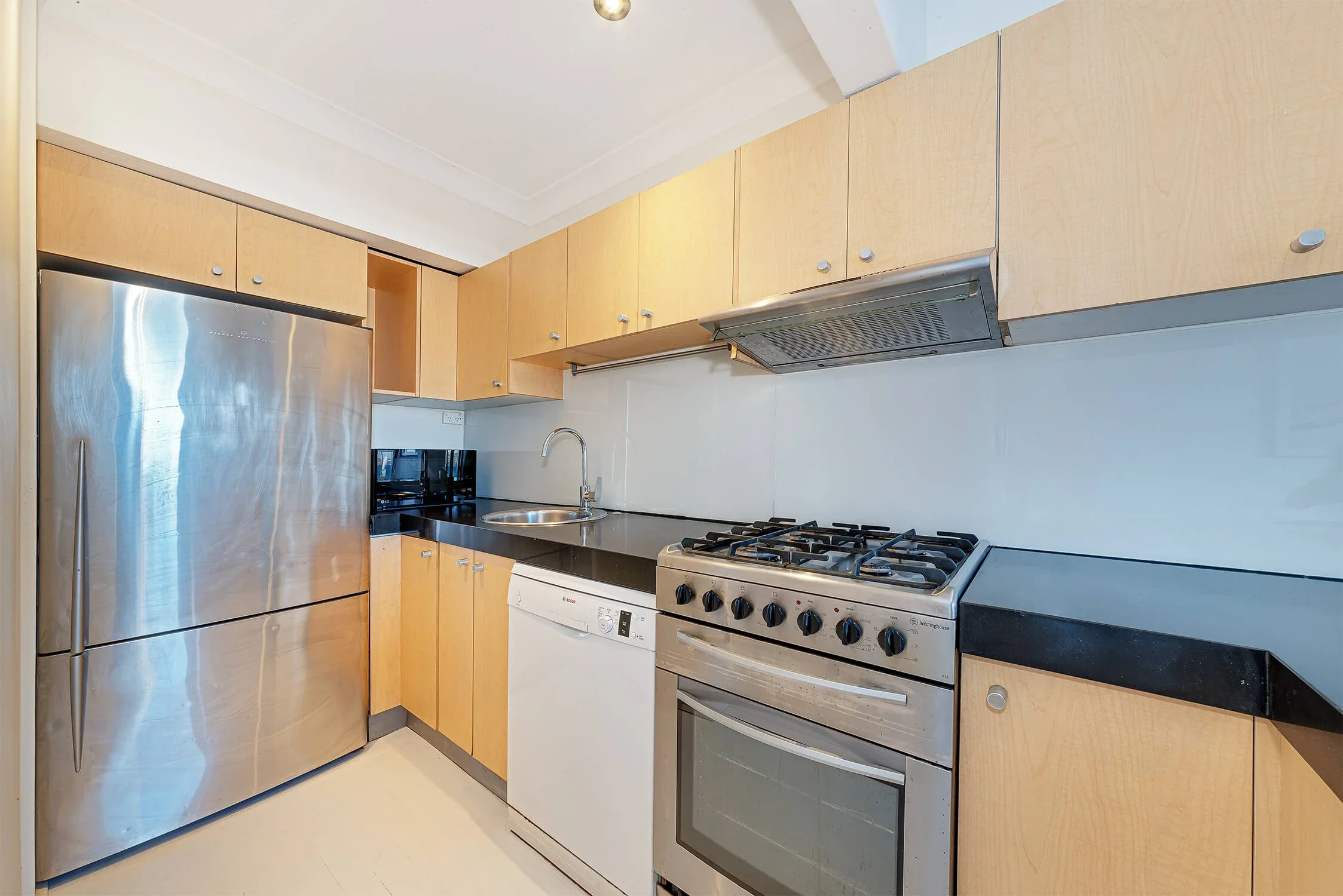 View of a modern kitchen with wooden cabinets, stainless steel refrigerator, oven, and dishwasher, along with a black countertop and a small sink.