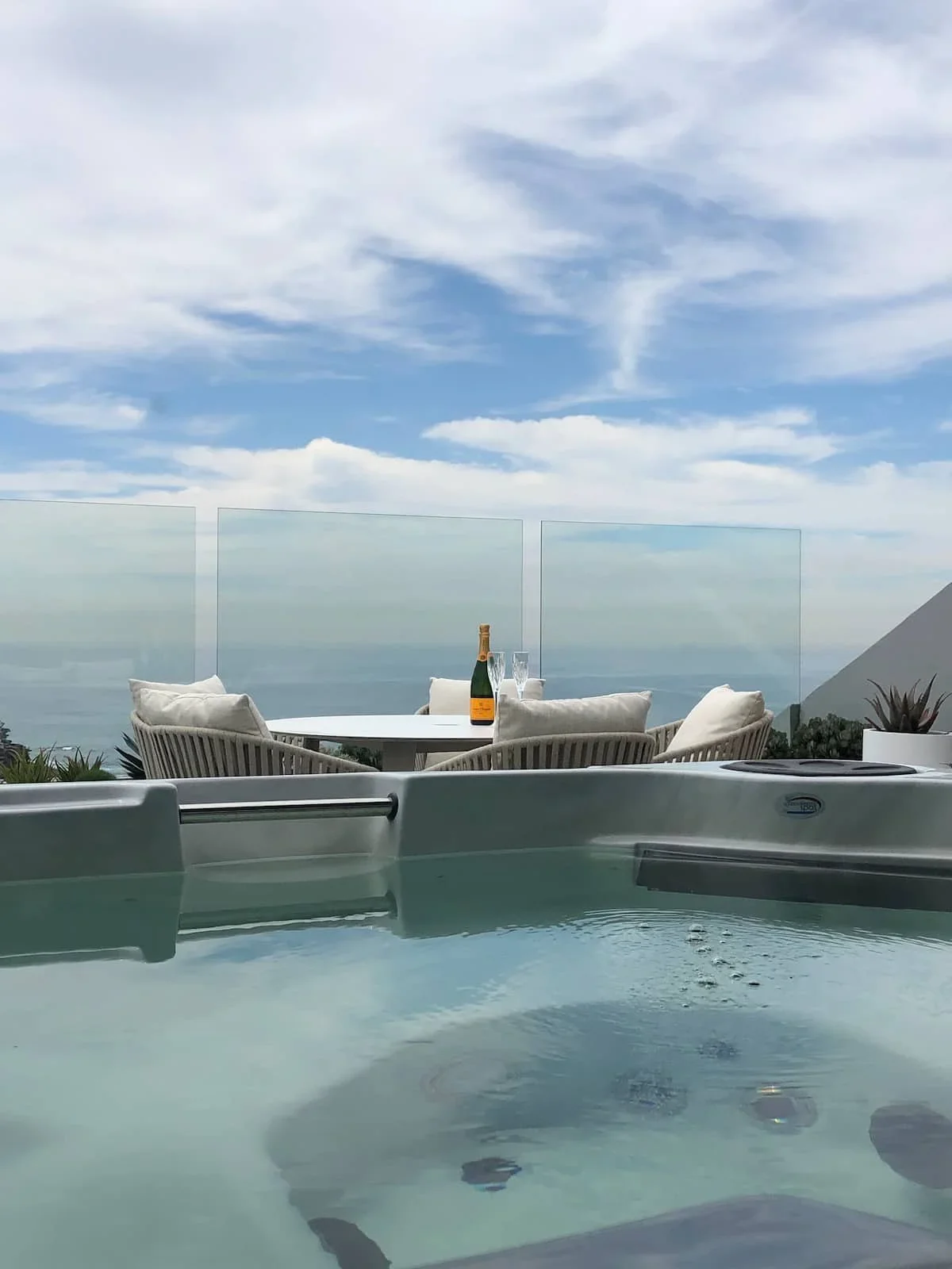 A view from a hot tub on a balcony with white chairs and a table set with a bottle of champagne and glasses, overlooking the ocean under a partly cloudy sky.