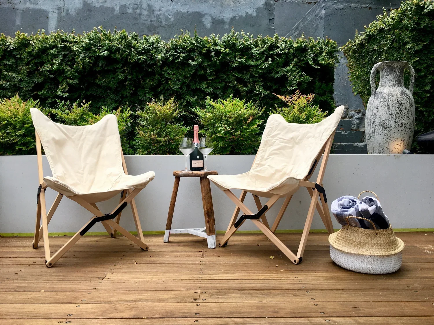 A cozy outdoor patio setup with two beige folding chairs, a small wooden table between them holding a bottle of wine and two wine glasses, a woven basket with a towel, and a large ceramic vase with a textured finish set against a lush green hedge.
