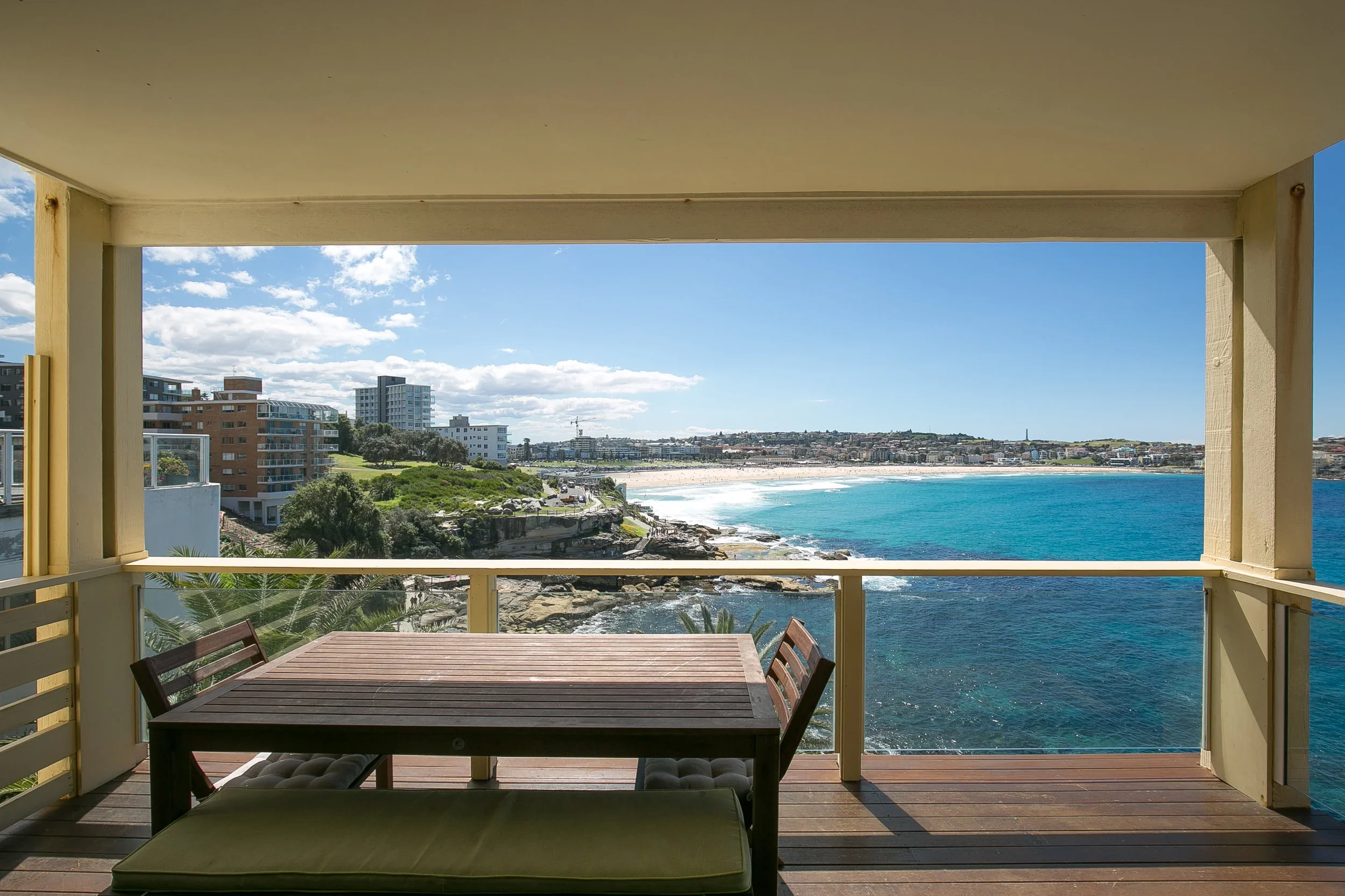 Balcony with a wooden table and chairs overlooking ocean and beach with buildings skyline in the distance under partly cloudy sky.