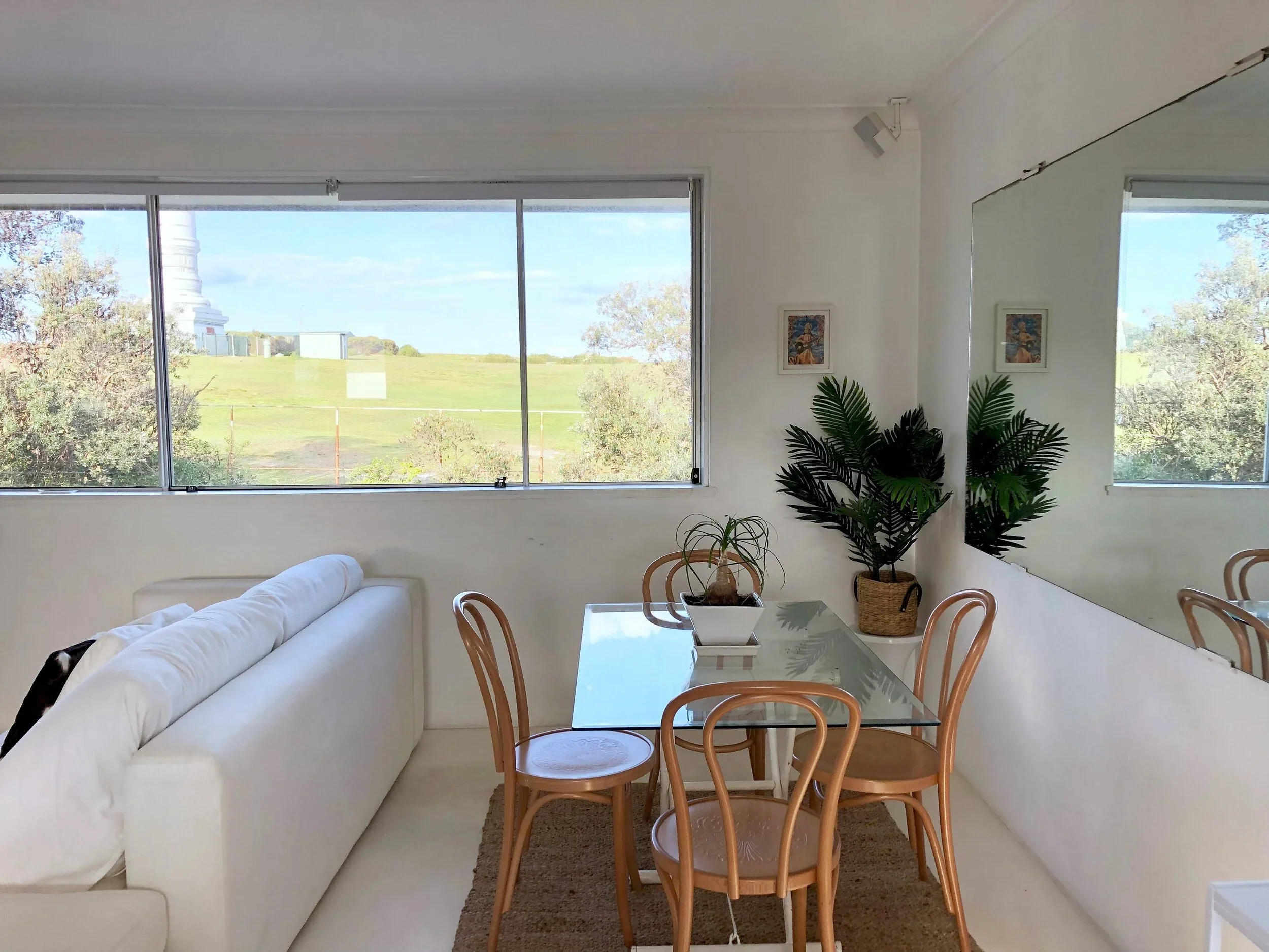 Living room with a white sofa, wooden dining table with chairs, large window with a view of greenery and a white monument outside, indoor plants, and mirror reflecting the room.