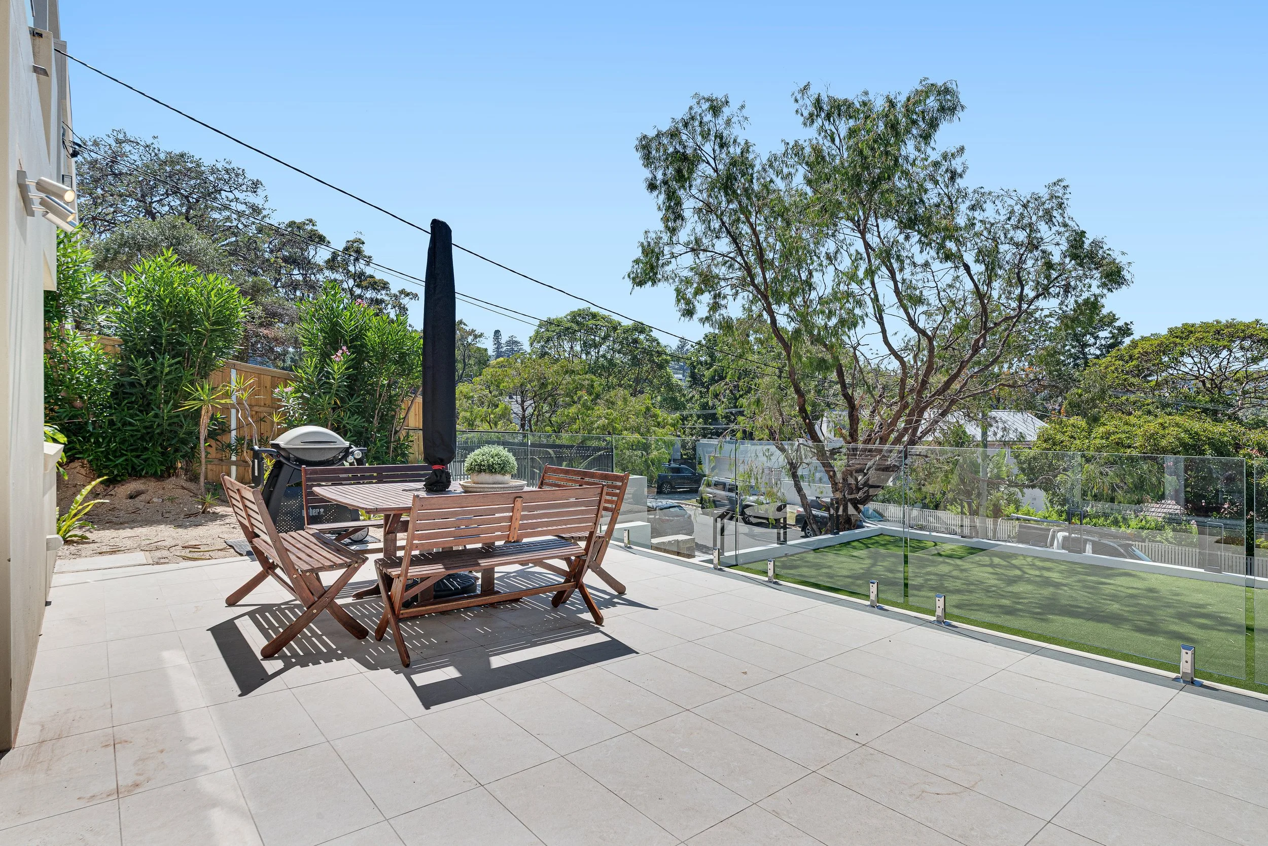 Patio area with wooden table and benches, surrounded by lush green trees and bushes, under a clear blue sky.