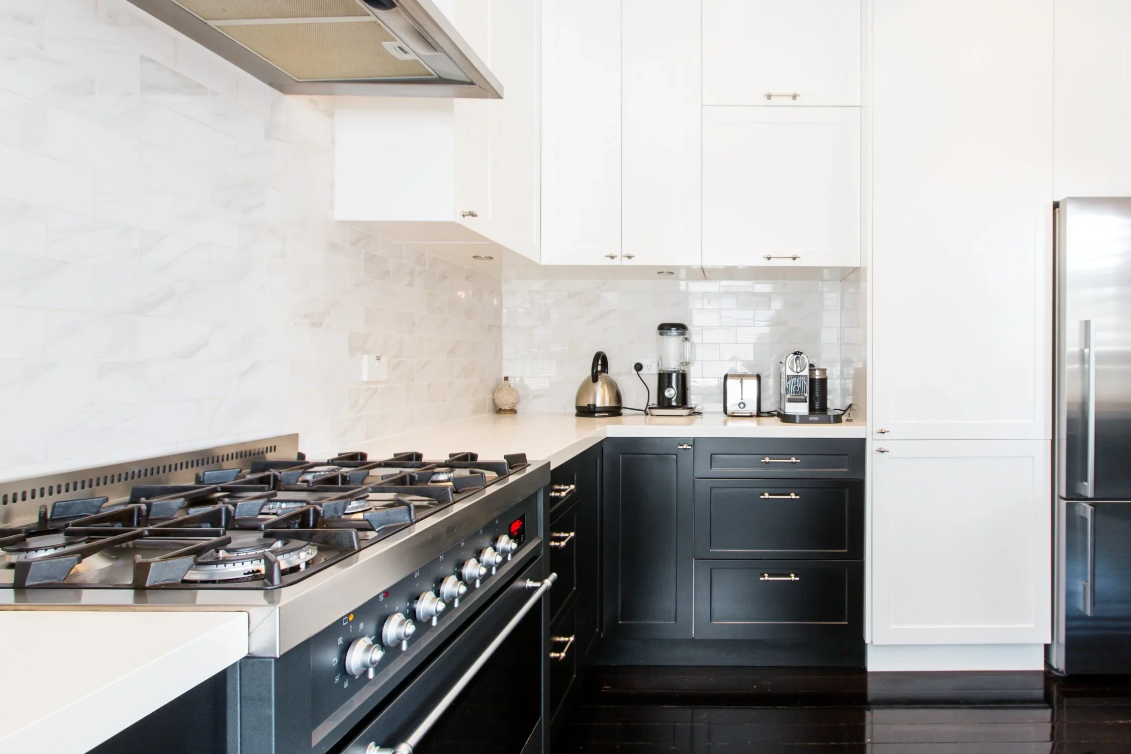 Modern kitchen with white and black cabinets, stainless steel stovetop, white countertops, and various small appliances such as a kettle, blender, and coffee maker.