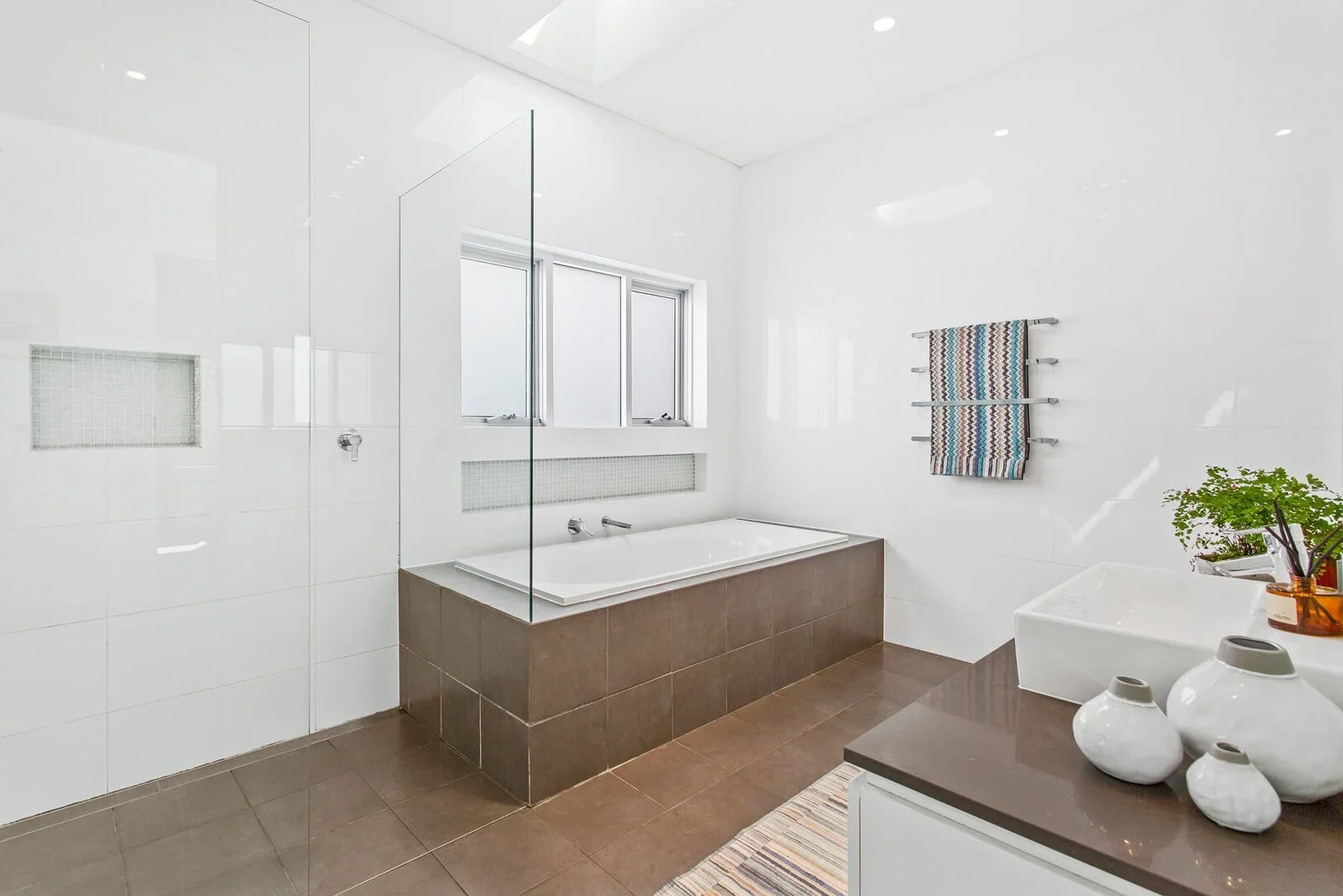 Modern bathroom with white tiled walls, a built-in bathtub, a window, a towel hanging on the wall, and a countertop with white vases and a plant.