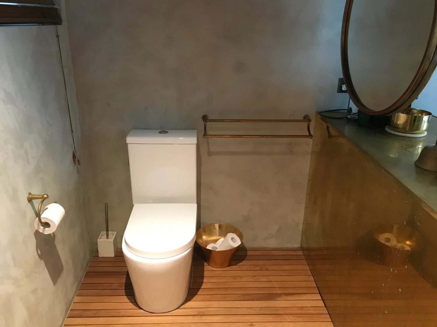 A modern bathroom with a white toilet, gold-colored toilet paper holder and waste bin, wooden flooring, a sink with a black countertop, a mirror, and a gold-colored bowl on the counter.