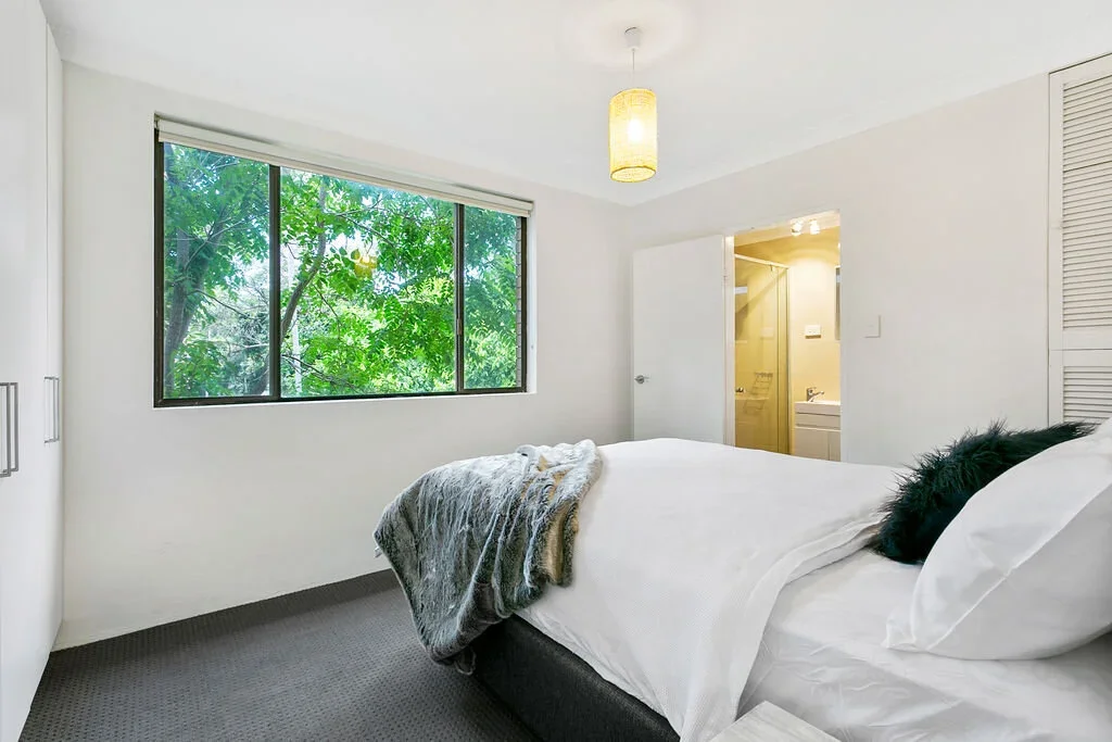 A bedroom with a bed, window overlooking green trees, and an open door leading to a bathroom.