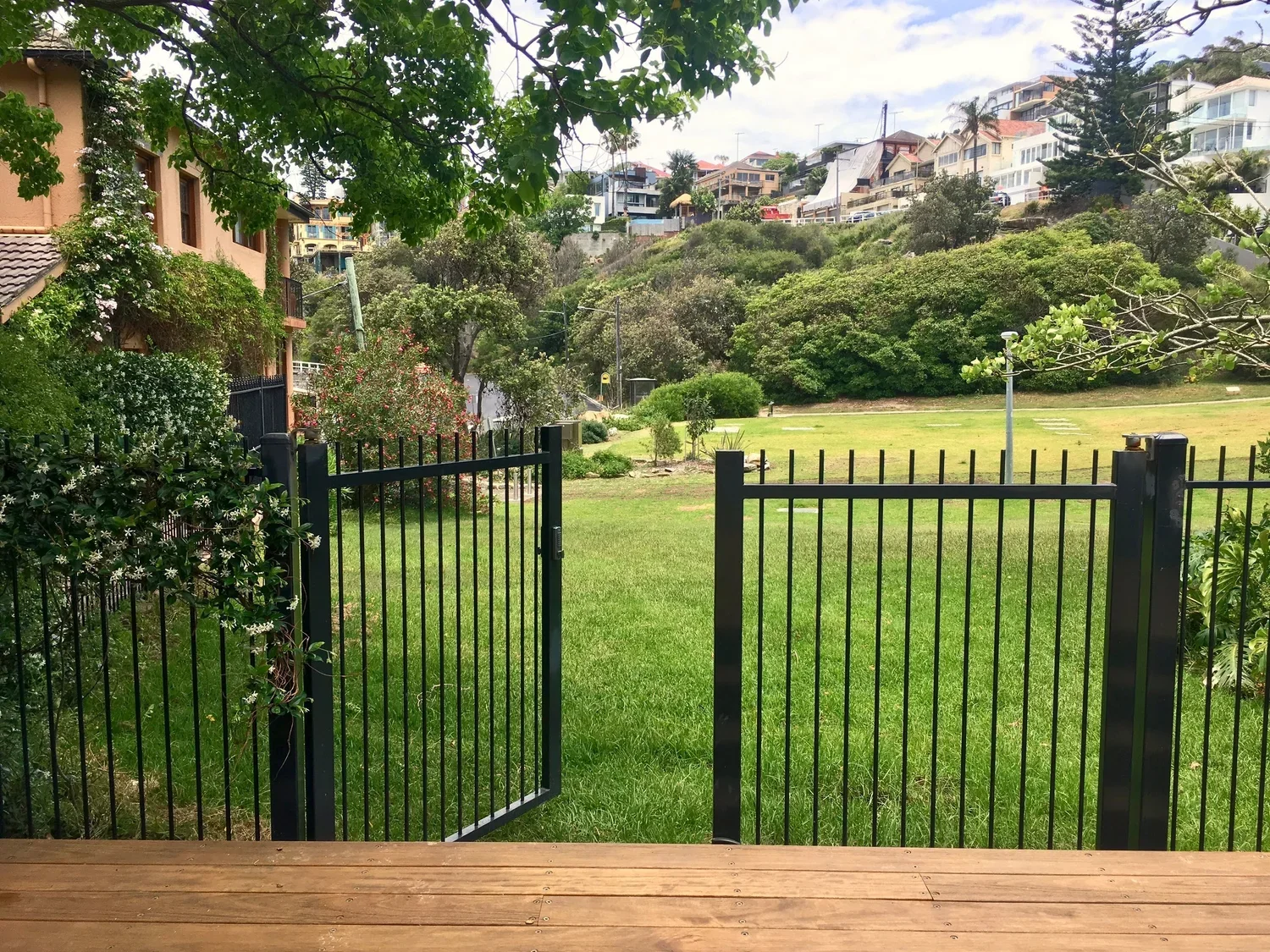 View of a fenced backyard with a grassy lawn, trees, bushes, houses on a hillside, and a partly cloudy sky.