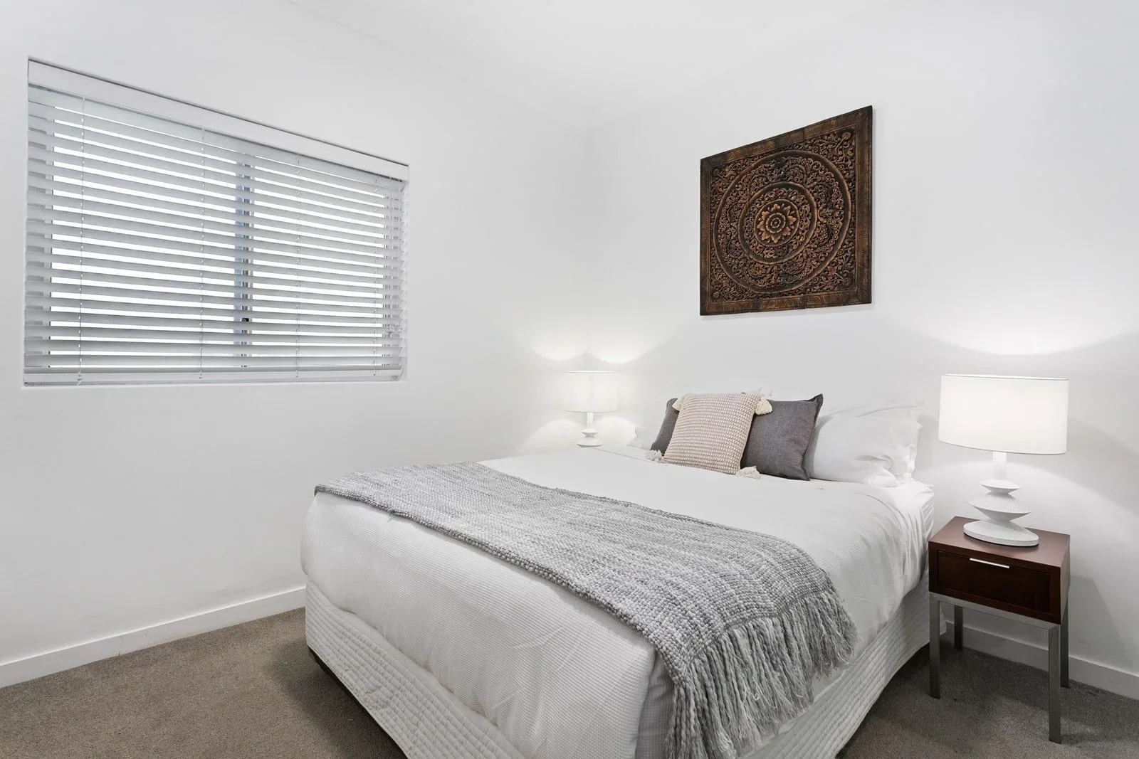 A bedroom with a white bed, grey and beige pillows, a grey throw blanket, white walls, a window with white blinds, a wooden nightstand, and a decorative wooden wall art piece.