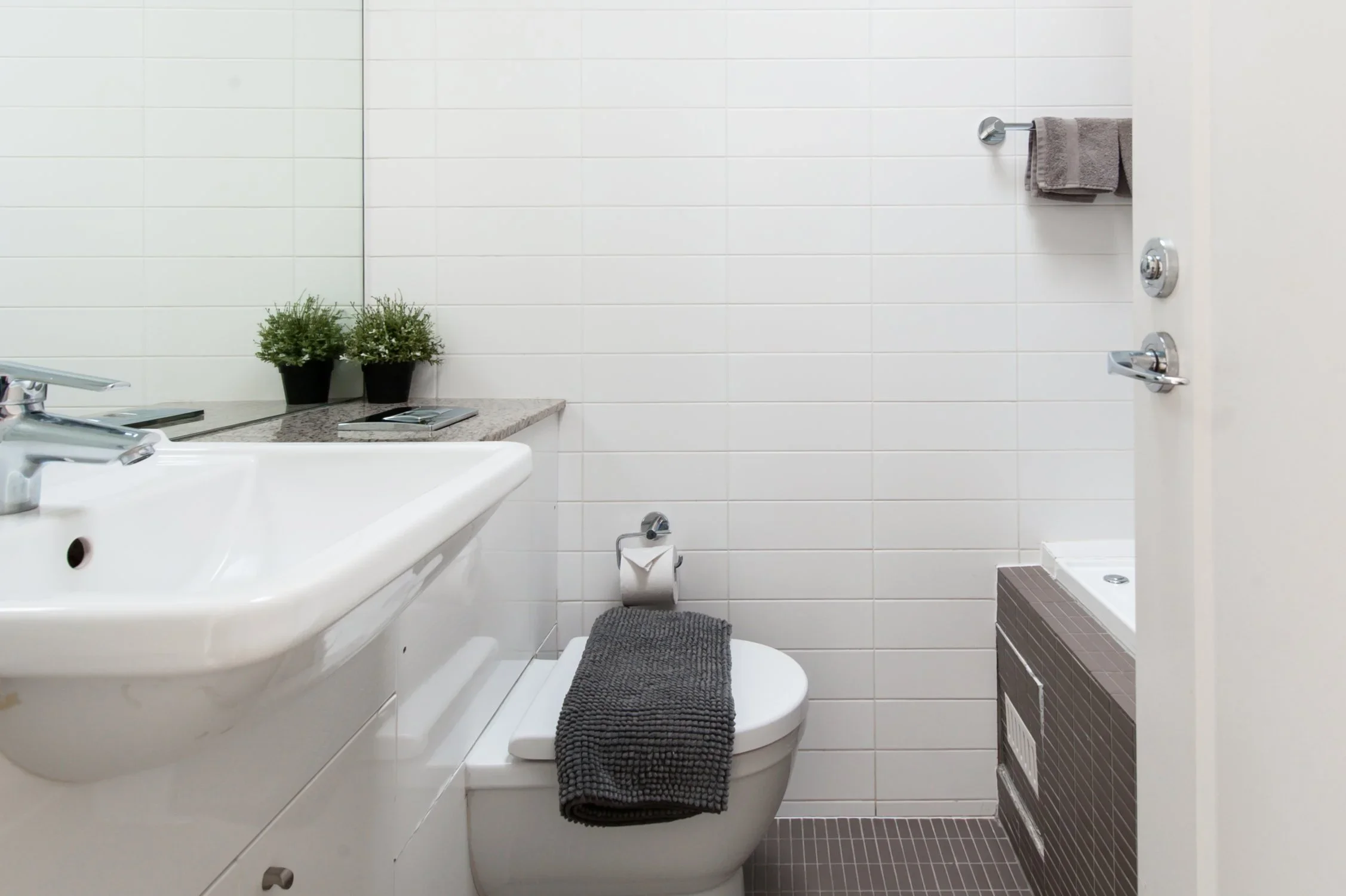 A modern bathroom with white tiled walls, a white sink, a toilet with a gray towel draped on it, a toilet paper holder, and a small shower area with a gray towel hanging on a hook.