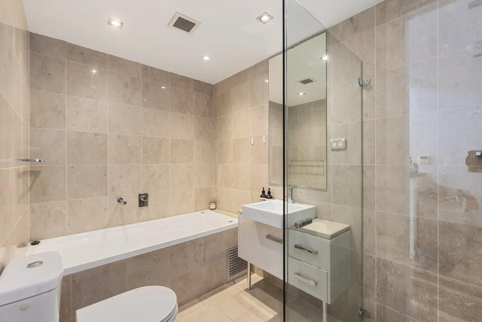 Modern bathroom with beige tiled walls, a bathtub, a small white sink with a mirror, and a glass shower enclosure.