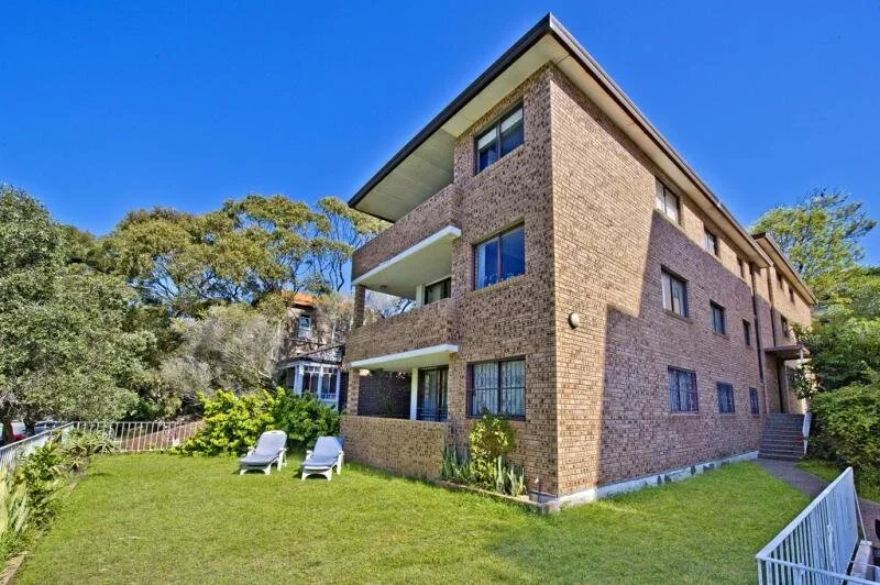 A three-story brick residential building with balconies and multiple windows, surrounded by a grassy yard with two lounge chairs and outdoor plants, under a clear blue sky.
