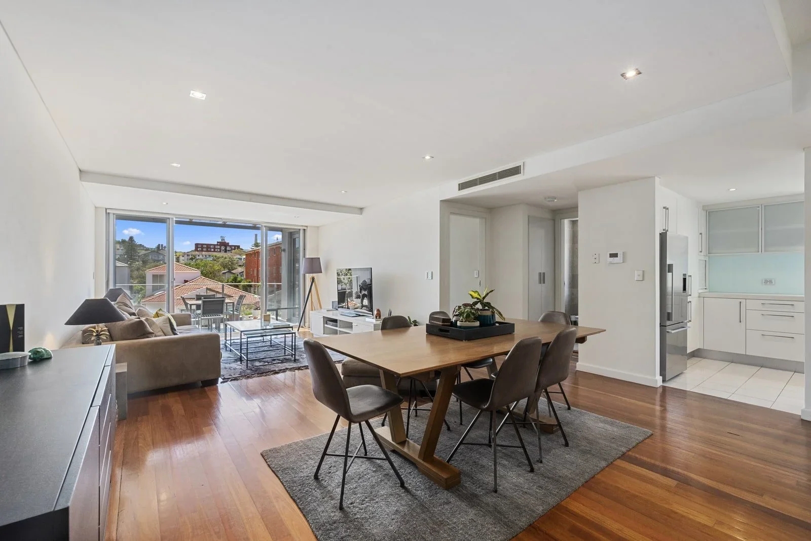 Open-concept living and dining area with wooden floors, large window, beige sofa, coffee table, TV, dining table with six chairs, and kitchen in background.