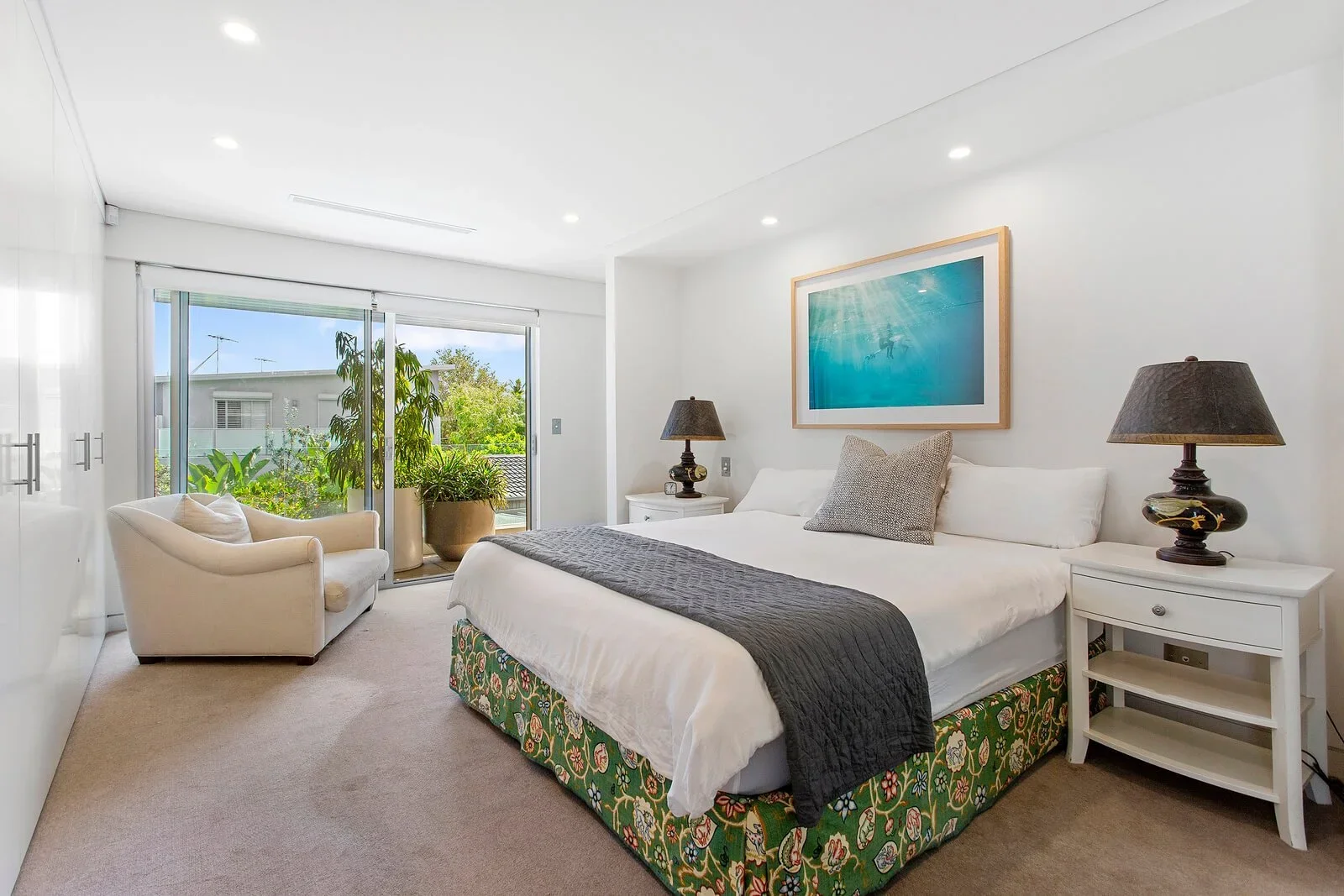 Bright bedroom with a large bed, nightstands with lamps, a white armchair near sliding glass doors, potted plants outside, and an underwater-themed artwork on the wall.