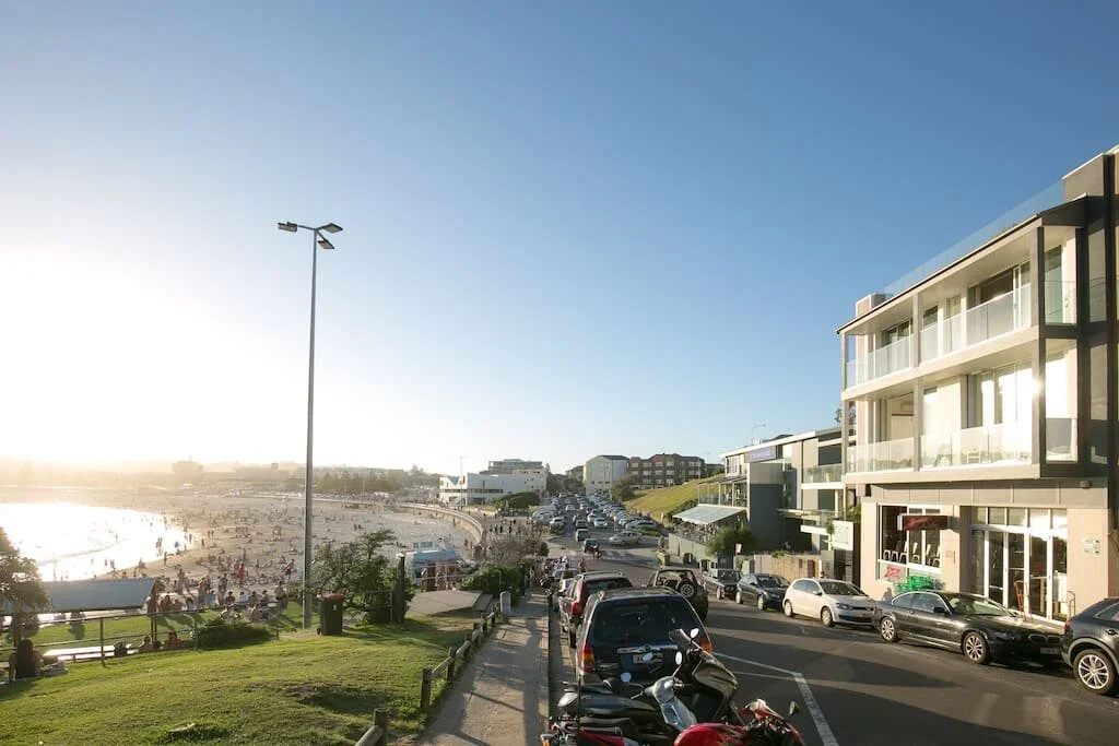 Beachside street with parked cars, modern apartments, and a busy sandy beach filled with people, near the water during daytime.