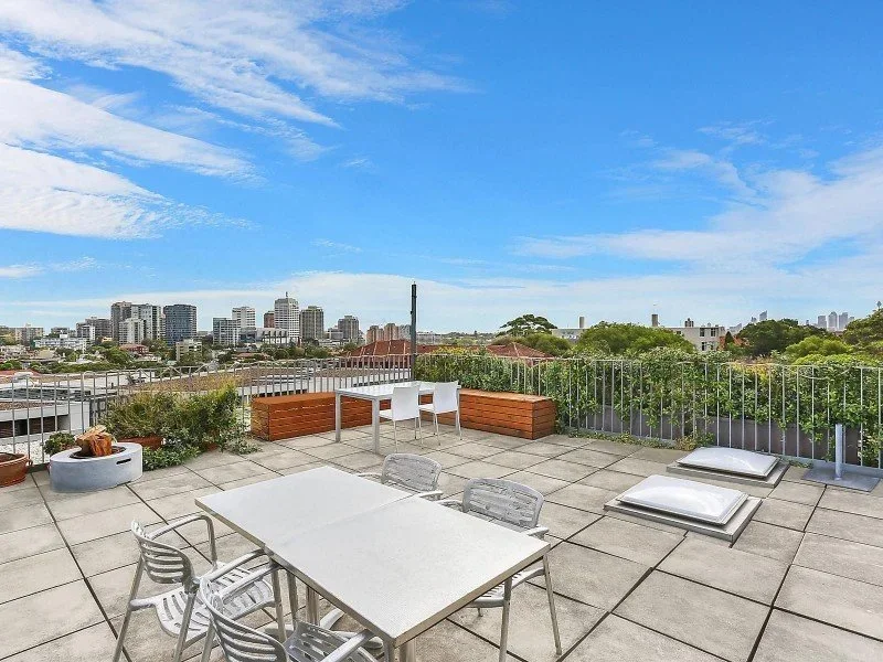 Rooftop patio with white table and chairs, potted plants, and city skyline view under a partly cloudy sky.