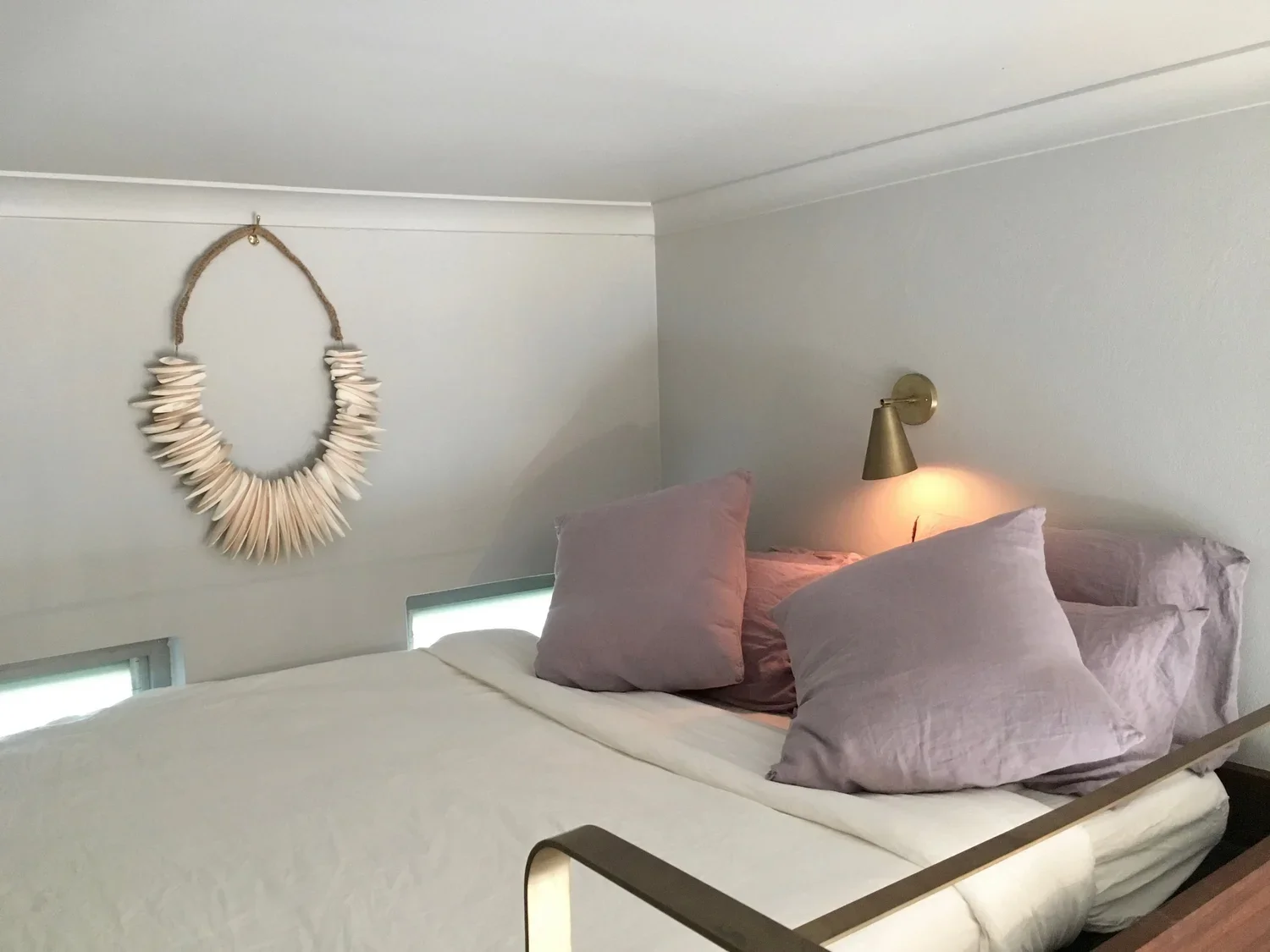 A cozy bedroom with a white bed, four pink pillows, a gold wall-mounted lamp, and a white shell necklace wall hanging.