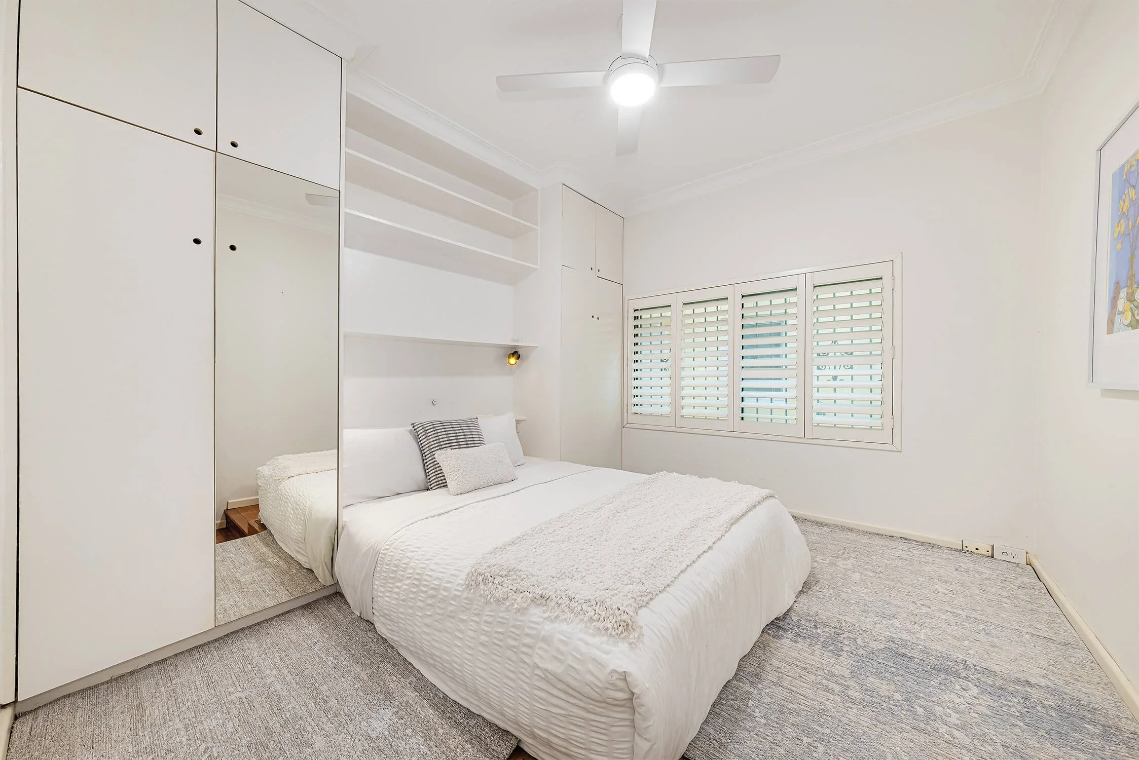 Minimalist bedroom with white walls, a bed with white bedding and pillows, a wardrobe with mirrors, a window with shutters, a ceiling fan, and artwork on the wall.