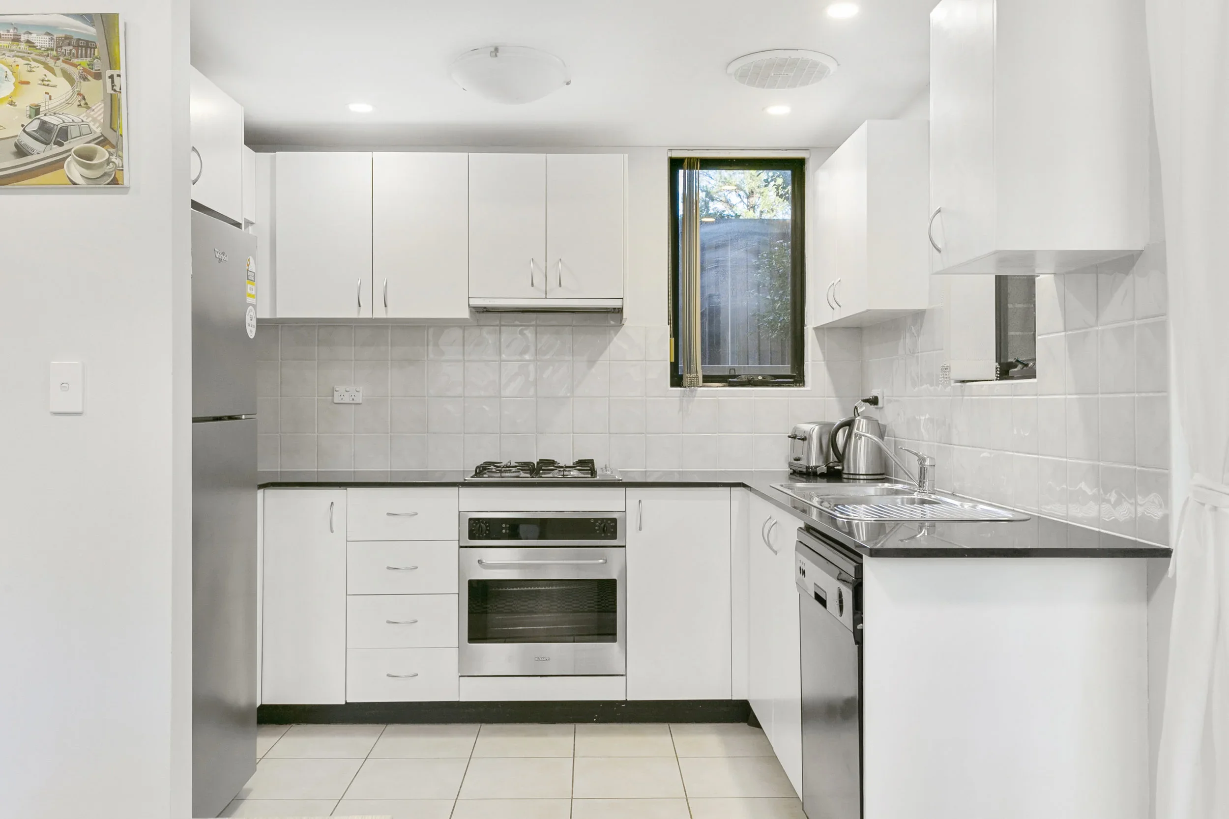 A compact white kitchen with upper and lower cabinets, a gas stove, oven, stainless steel refrigerator, and small appliances on the counter, including a toaster, kettle, and coffee maker, with a window above the sink.