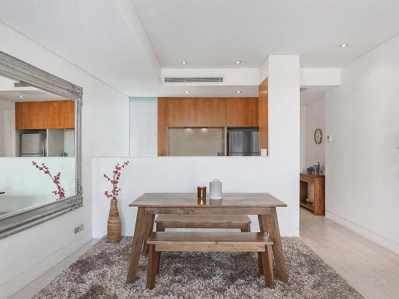 Minimalist dining area with a wooden table, matching bench, and decorative vases, with a kitchen in the background.