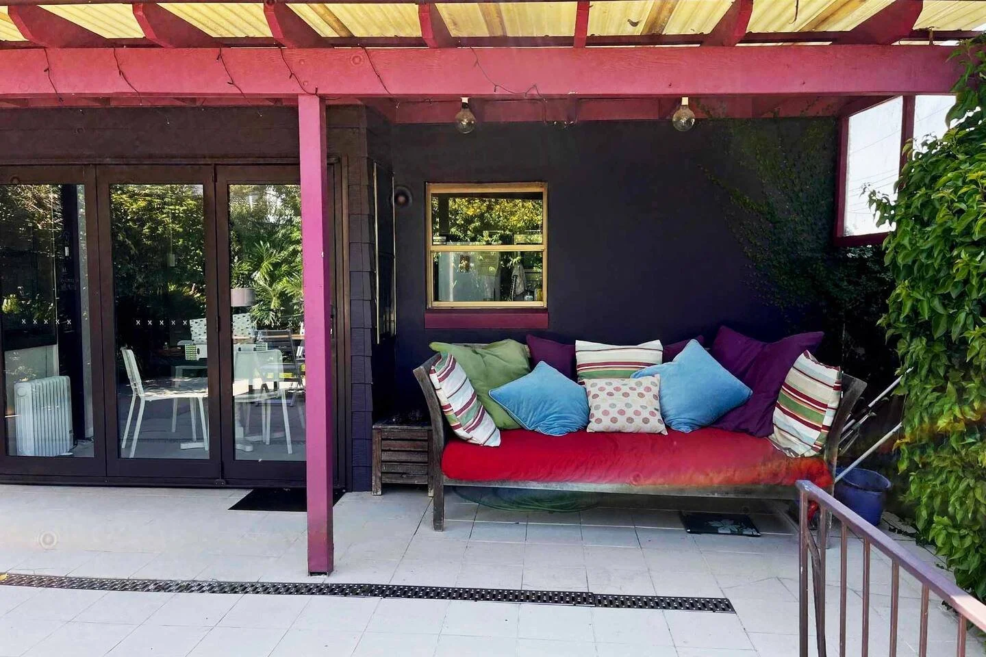 An outdoor patio area with a cushioned bench and colorful pillows, adjacent to glass sliding doors leading inside a house, with a pink and black painted wall and greenery.