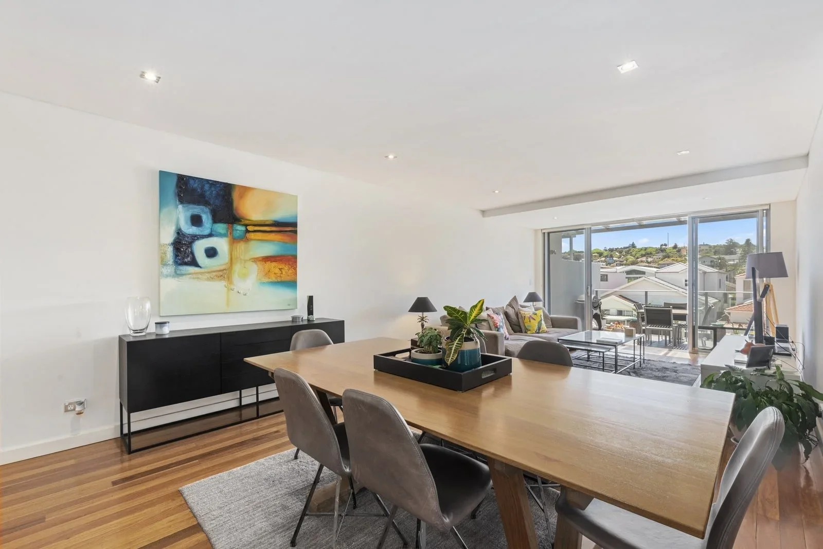 Modern living and dining room with large wooden table, gray chairs, black sideboard, abstract painting, sofa, coffee table, and sliding glass doors leading to a balcony with outdoor seating.