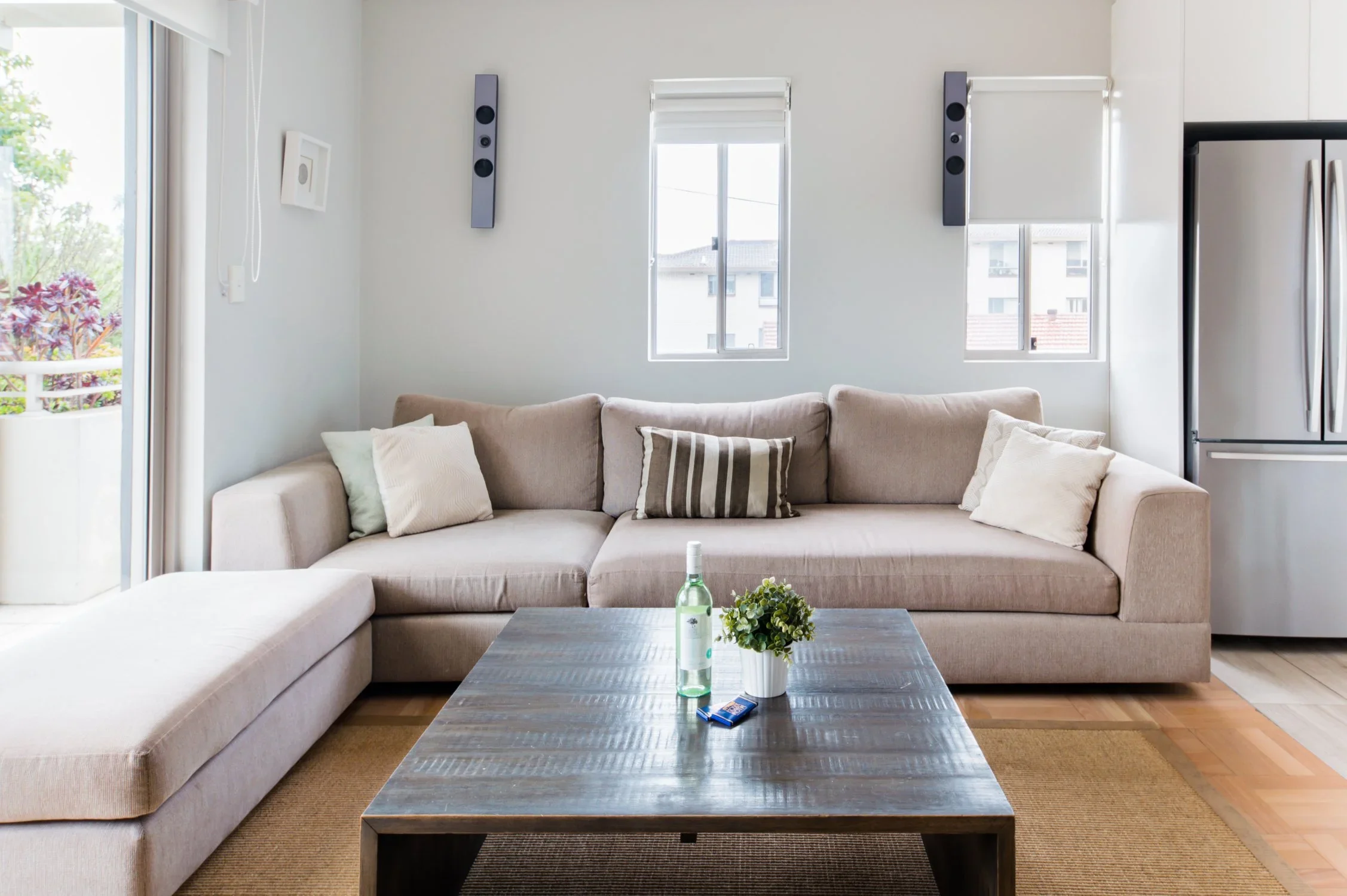 Living room with a beige sectional sofa, pillows, a wooden coffee table with a plant and bottle, two windows, and a refrigerator, in a bright space with hardwood and rug flooring.