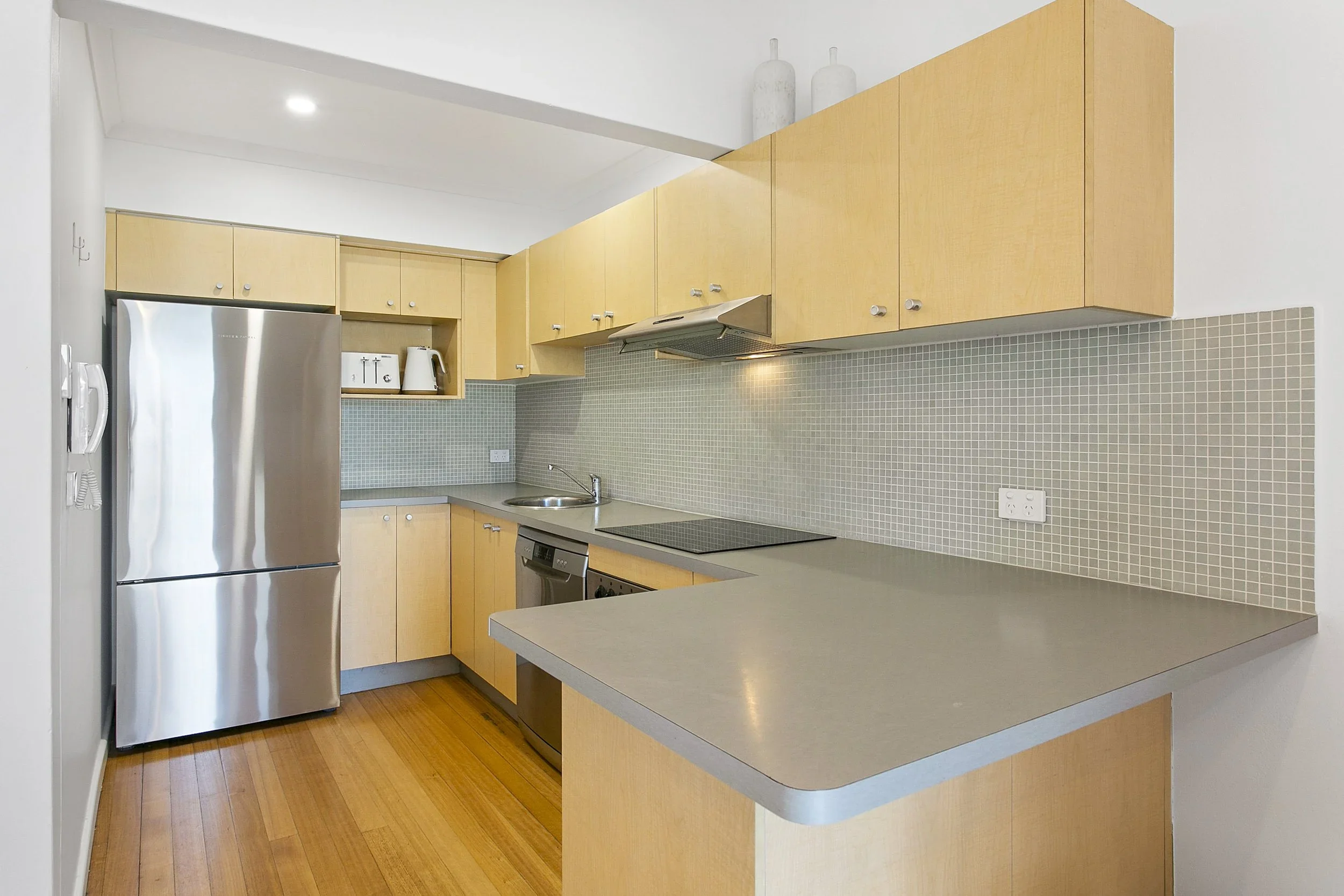 Modern kitchen with light wood cabinets, stainless steel refrigerator, microwave, stove, and dishwasher, with small mosaic tile backsplash and hardwood flooring.