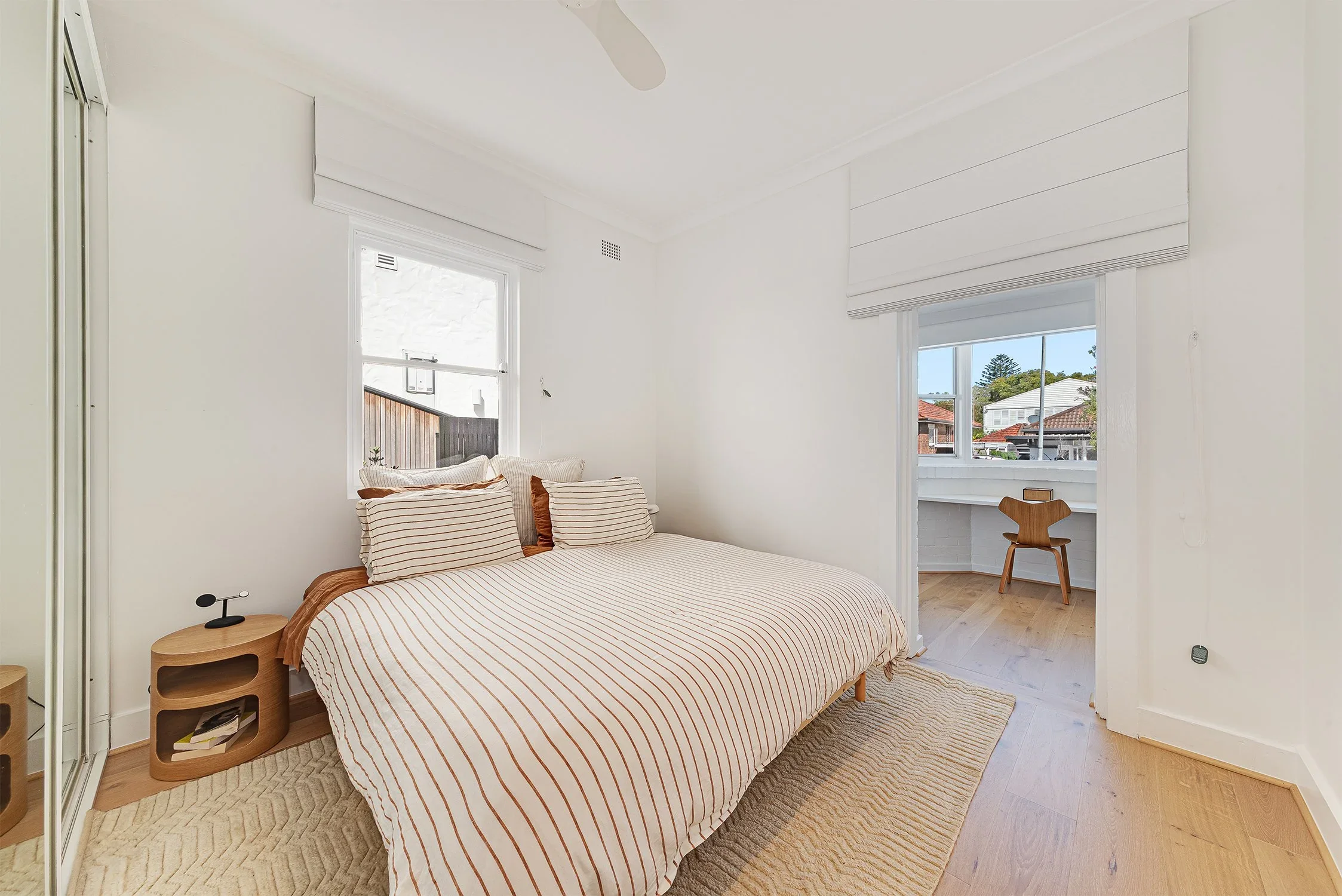 A bedroom with white walls, a bed with striped bedding, a small wooden nightstand, and a window with a view of neighboring houses and trees.