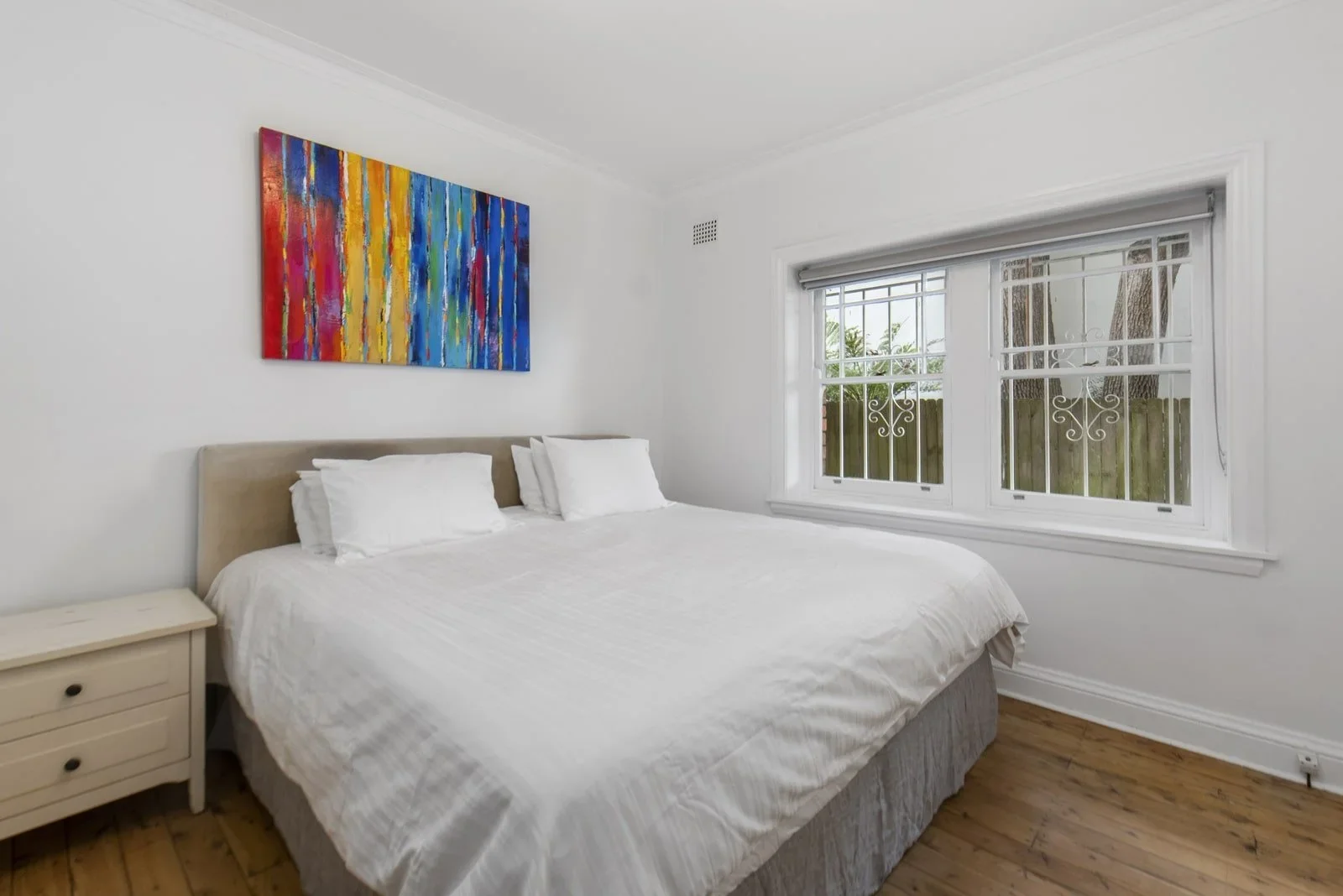 A bedroom with white walls, a bed with white bedding, two pillows, a beige headboard, a nightstand, a colorful abstract painting above the bed, and a window with white frames and decorative grilles showing greenery outside.