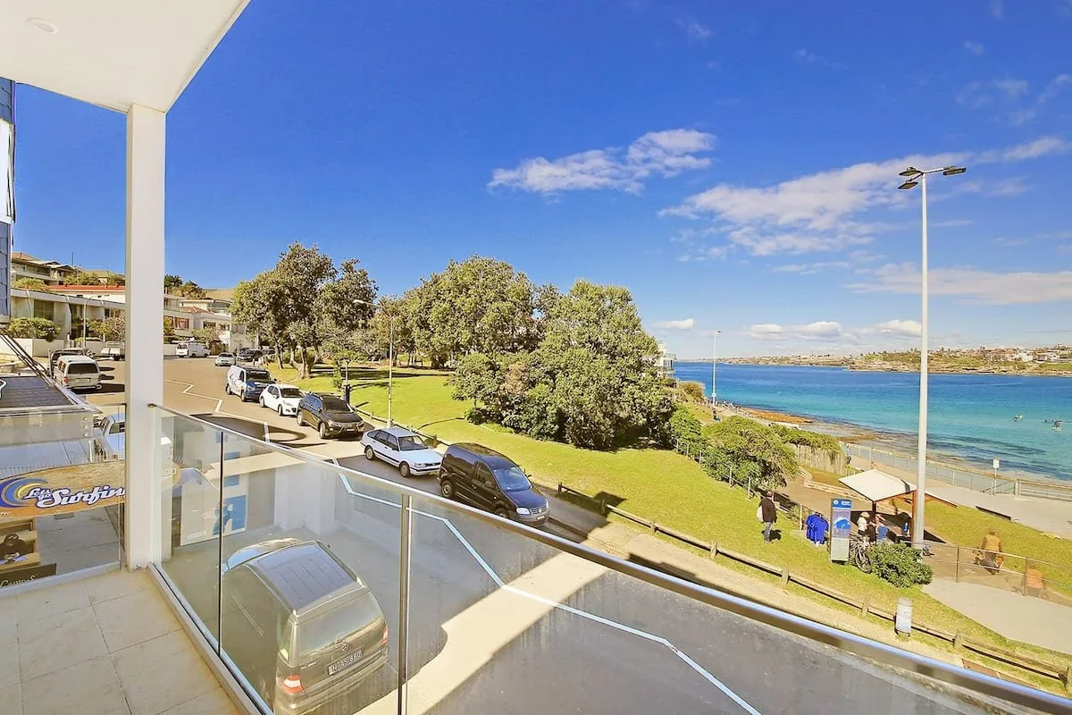 View from a balcony showing a parking lot with cars, a grassy area with trees, a beach, and the ocean under a blue sky with some clouds.