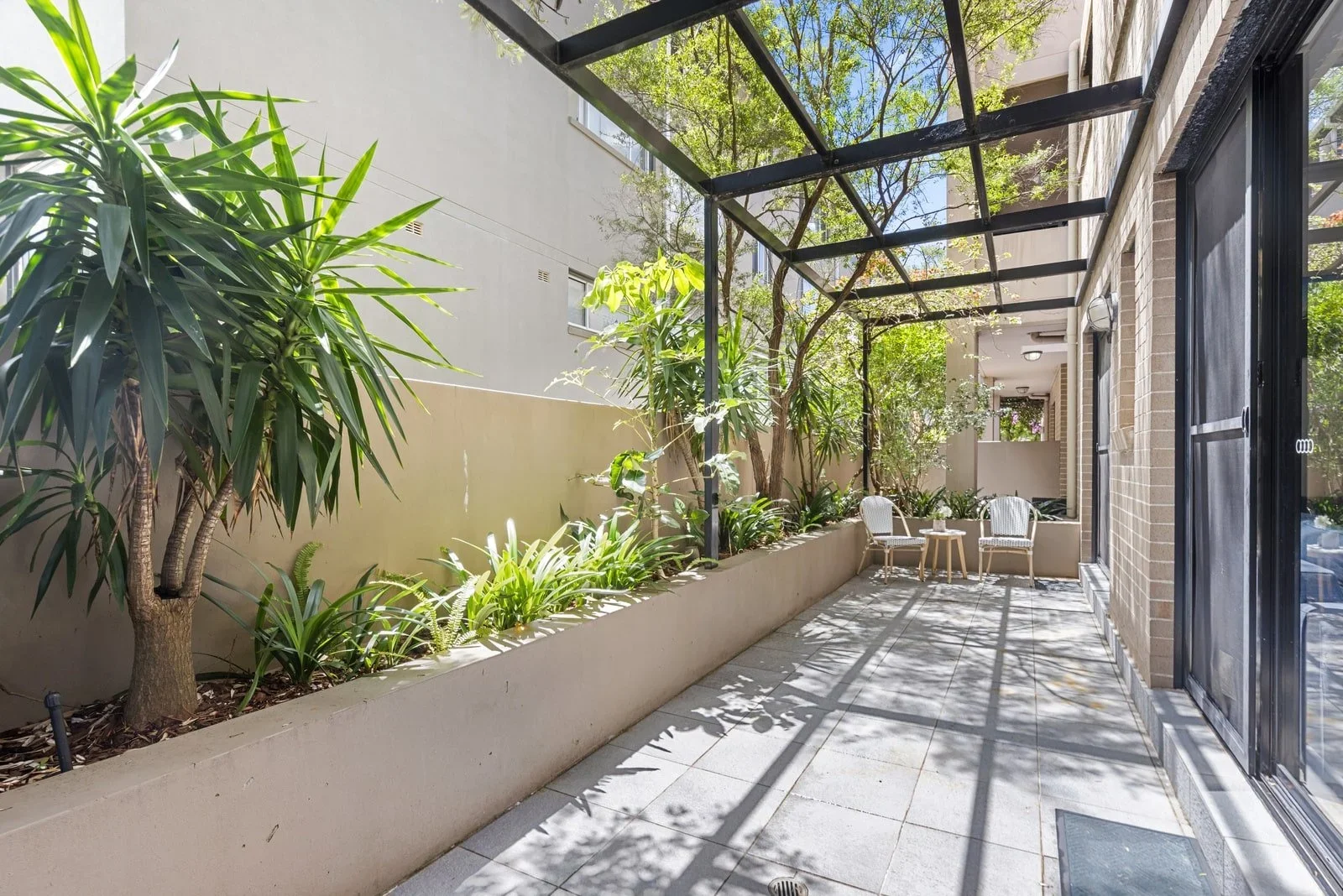 A sunny outdoor patio with a garden bed containing green plants and trees, patio furniture including two chairs and a small table, and a black metal framed glass roof providing partial shade.