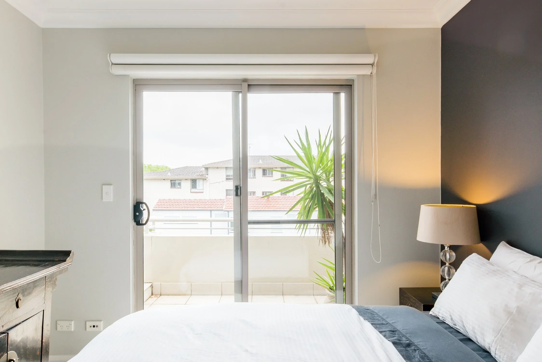 Bedroom with sliding glass door leading to balcony, view of neighboring buildings, bed with white linens, bedside lamp, and decorative plant outside.