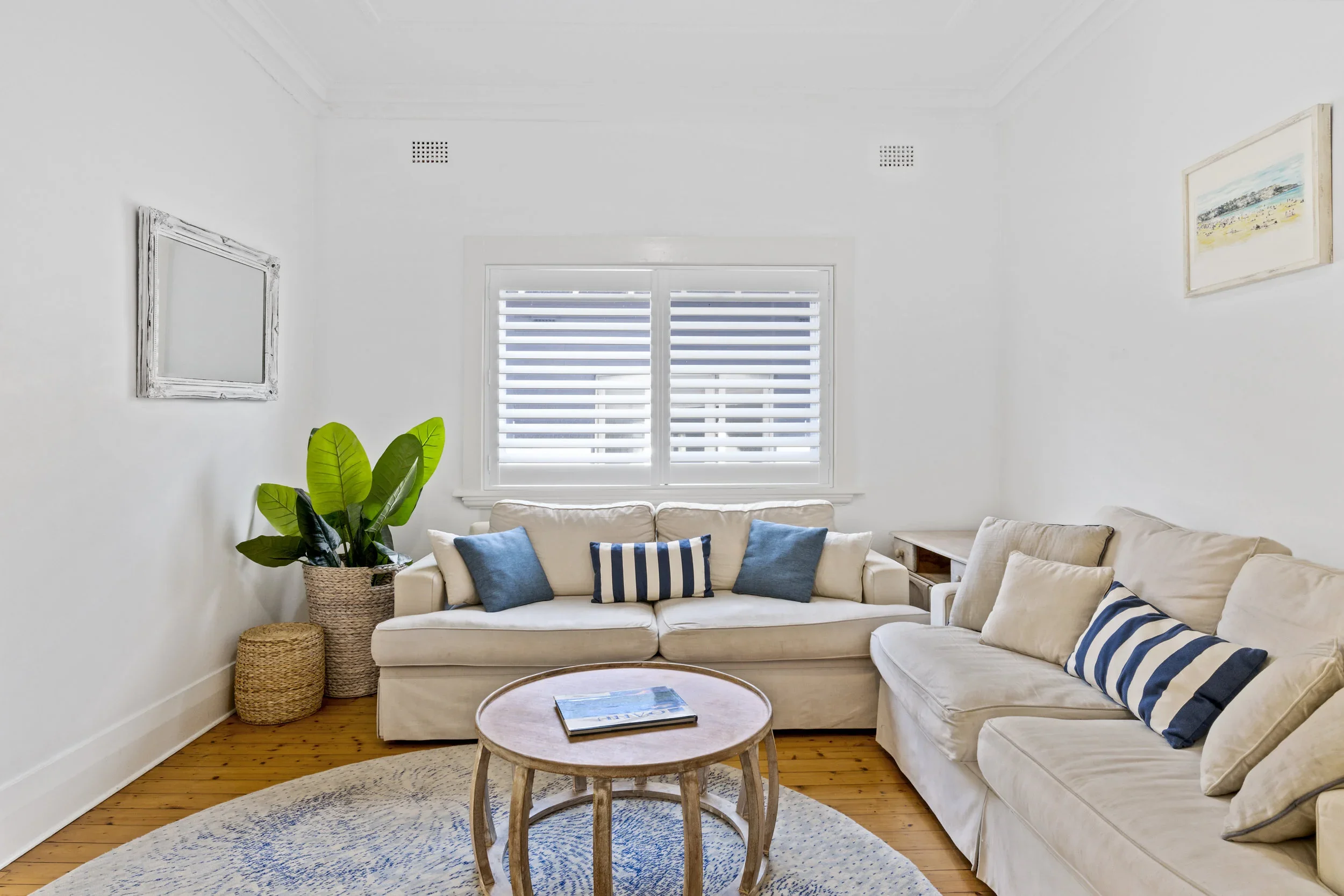 Bright living room with white walls, beige sofas with blue and white striped pillows, a round wooden coffee table, large window with white blinds, potted plant, framed wall art, wooden floor, and a patterned area rug.