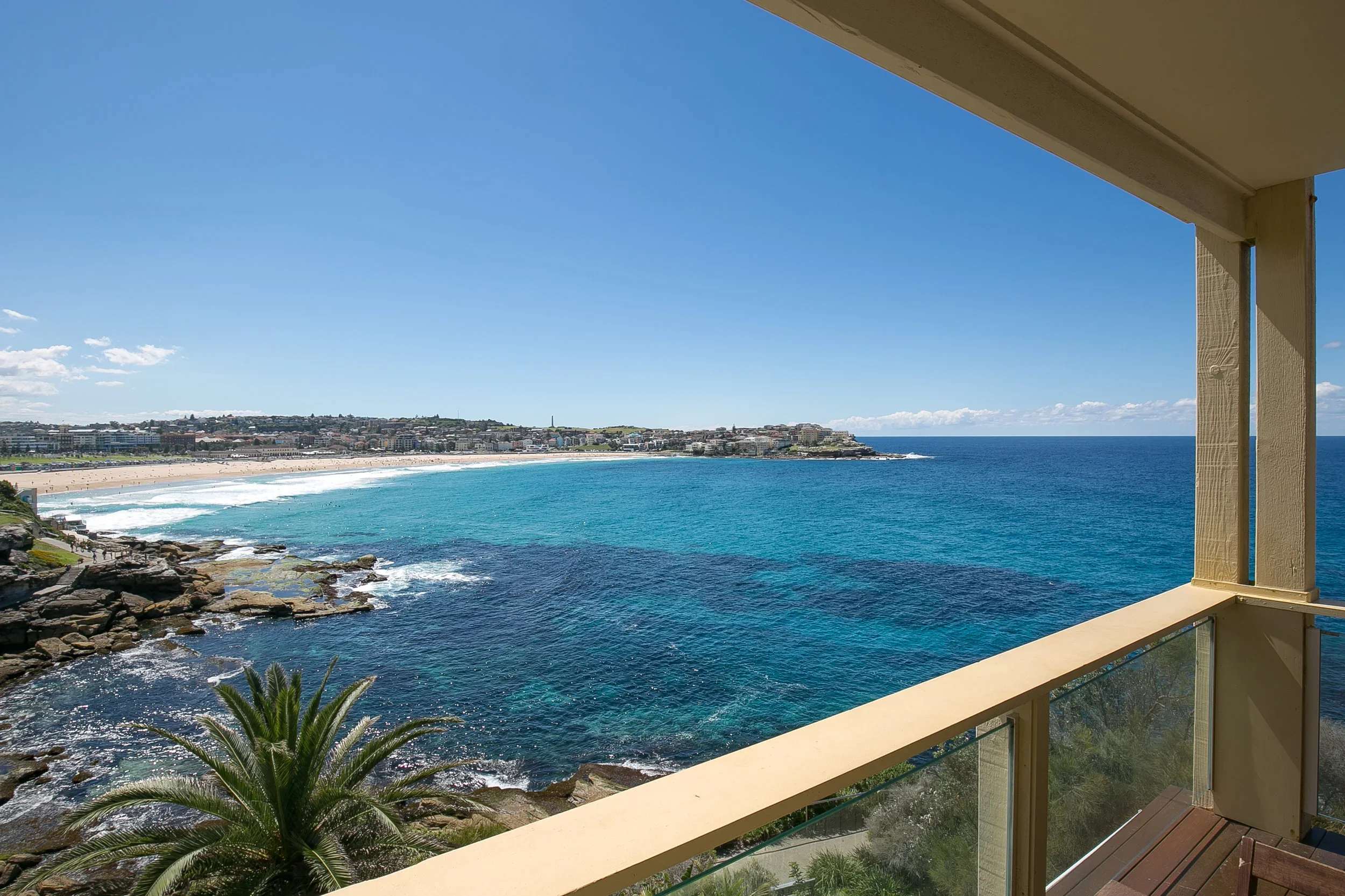 View of a beach and ocean from a balcony, with a clear blue sky and some clouds in the distance.
