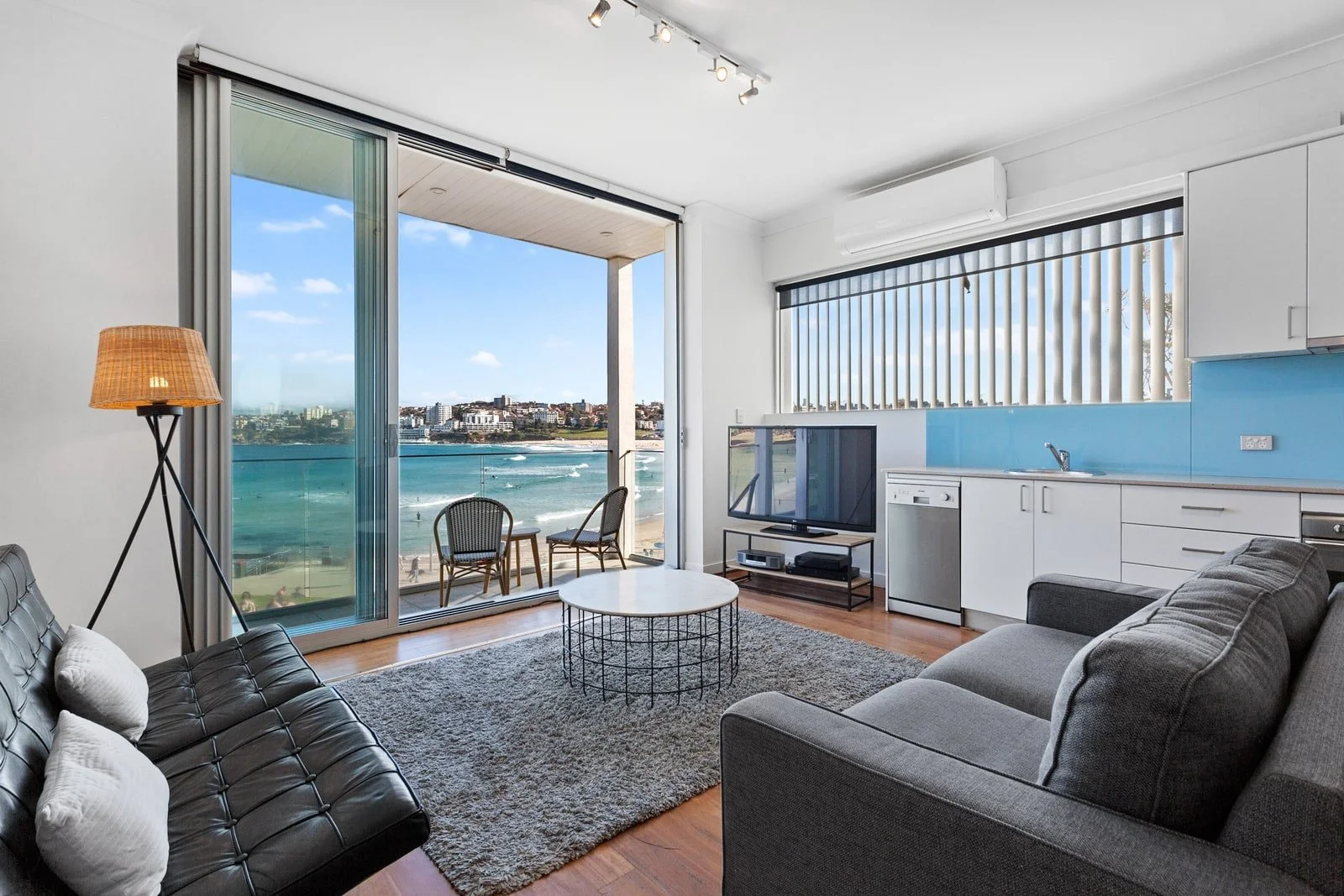 Living room with large window and sliding glass door revealing a beach view, containing a gray sofa, black leather couch, white coffee table, TV stand with television, blue kitchen backsplash, and small balcony with two chairs.